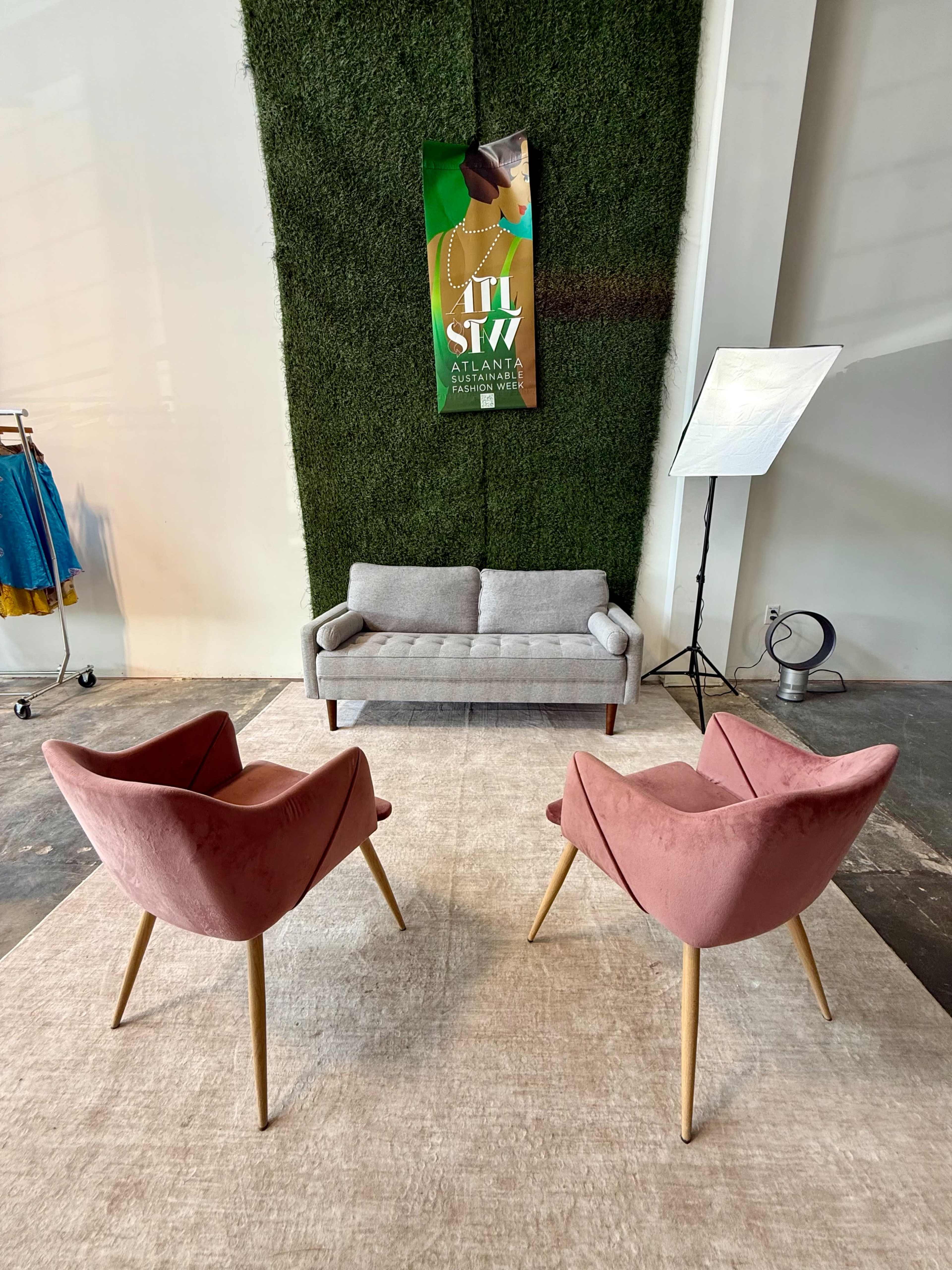 The image shows a minimalist seating area with a gray sofa and two pink armchairs arranged on a light rug against a green wall featuring a poster.