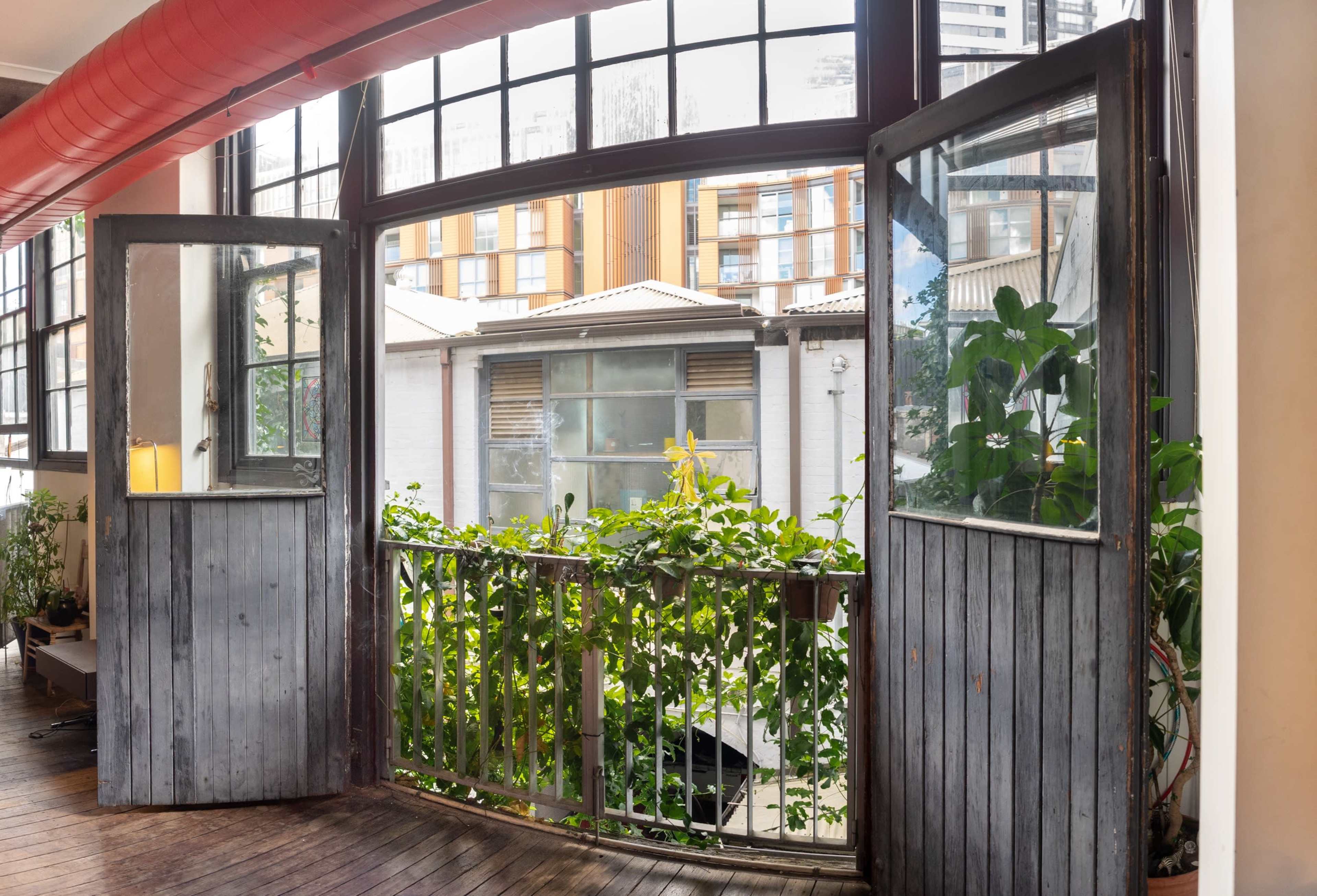 An open-segmented window with wooden frames overlooking a balcony filled with greenery and a view of nearby buildings.