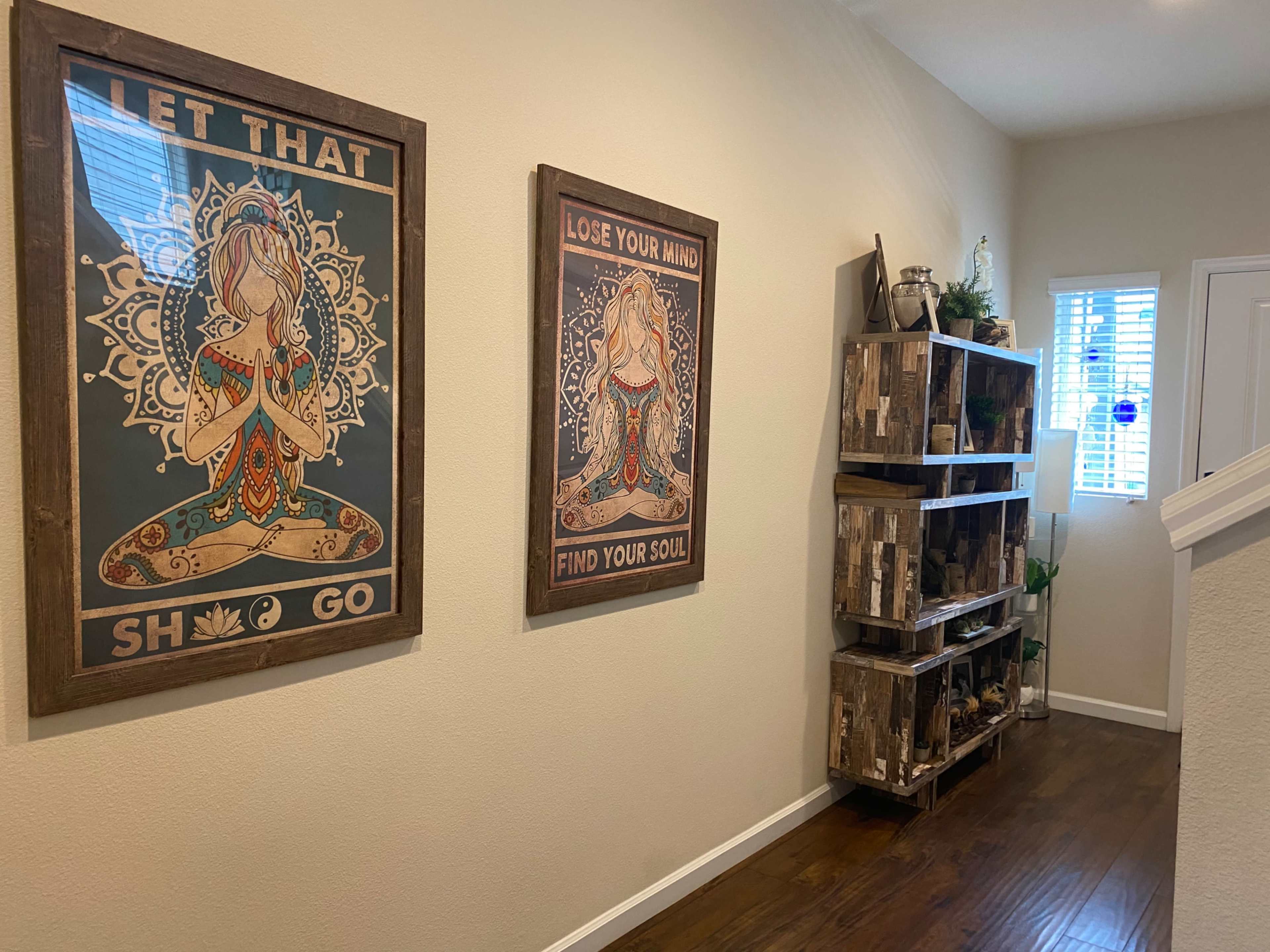 A hallway with two framed wall art pieces featuring meditative figures, and a wooden shelving unit displaying plants and decorative items.