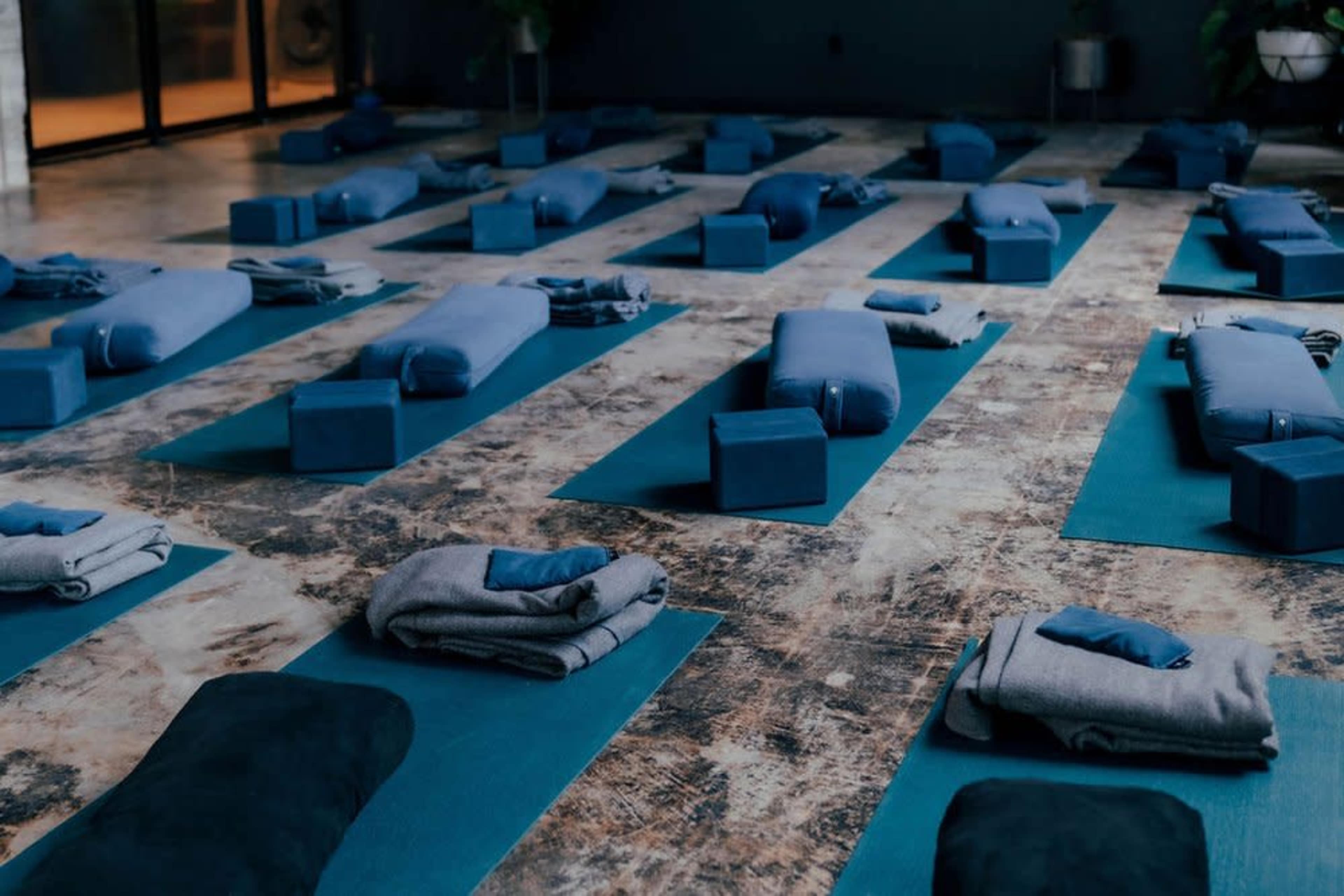 The image shows a yoga studio with neatly arranged blue mats, bolsters, and blocks on a textured floor.