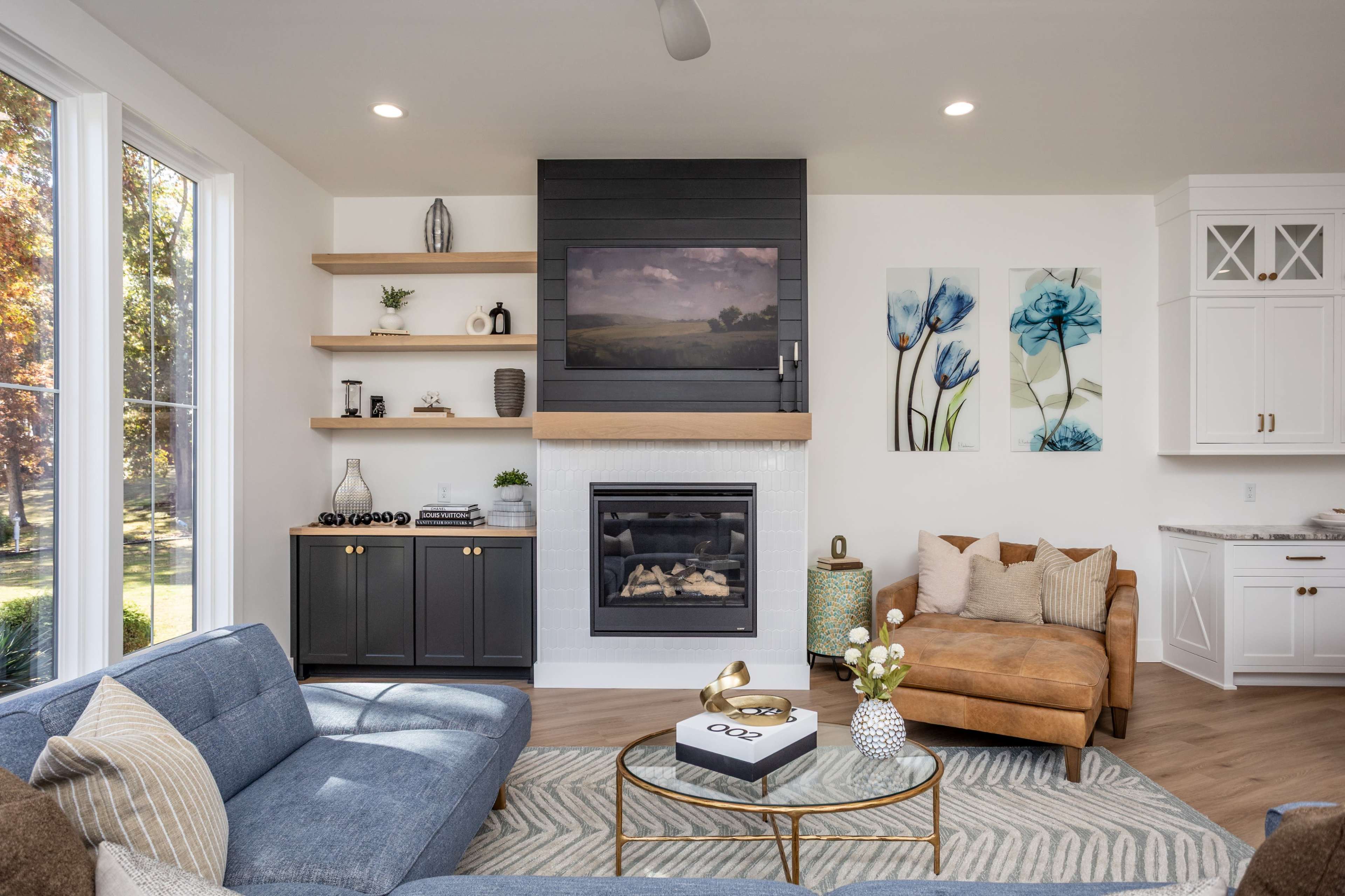 A modern living room features a blue sectional sofa, an orange armchair, a television mounted above a fireplace, and decorative shelves displaying various items.