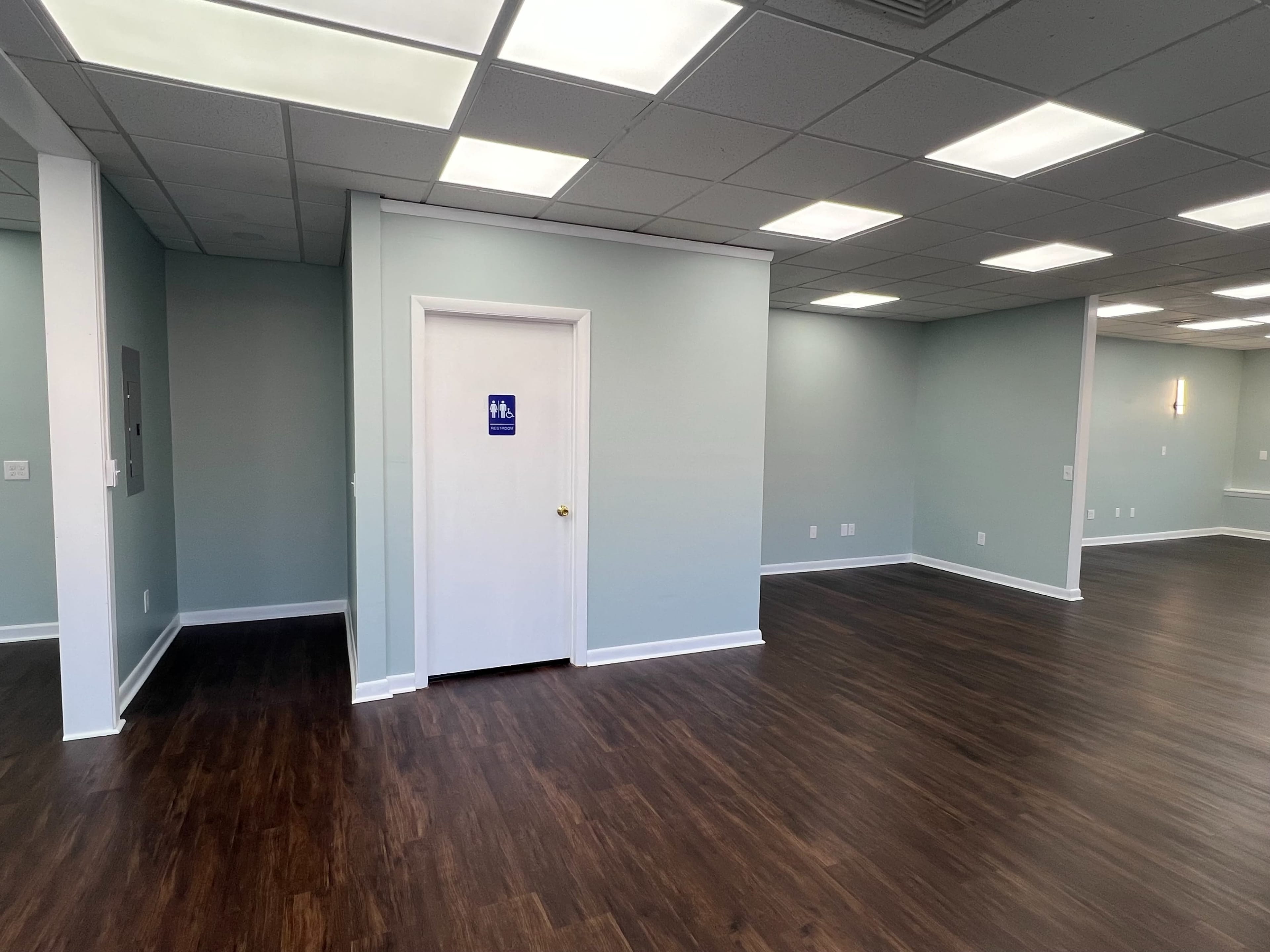The image shows an empty room with light blue walls, wooden flooring, and a door marked for restrooms.