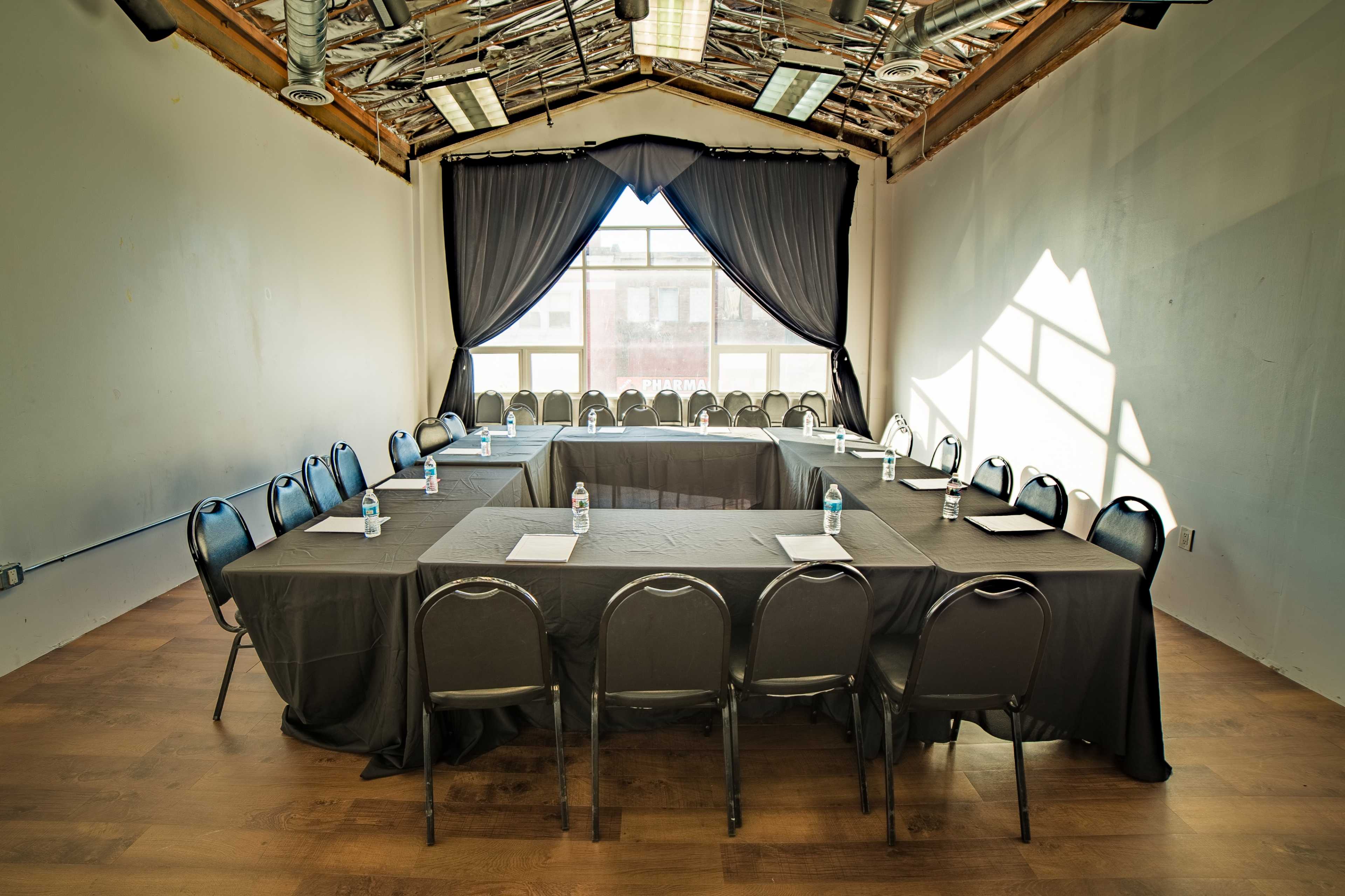 A conference room features two large tables arranged in a U-shape, surrounded by black chairs, with large windows allowing natural light to enter.