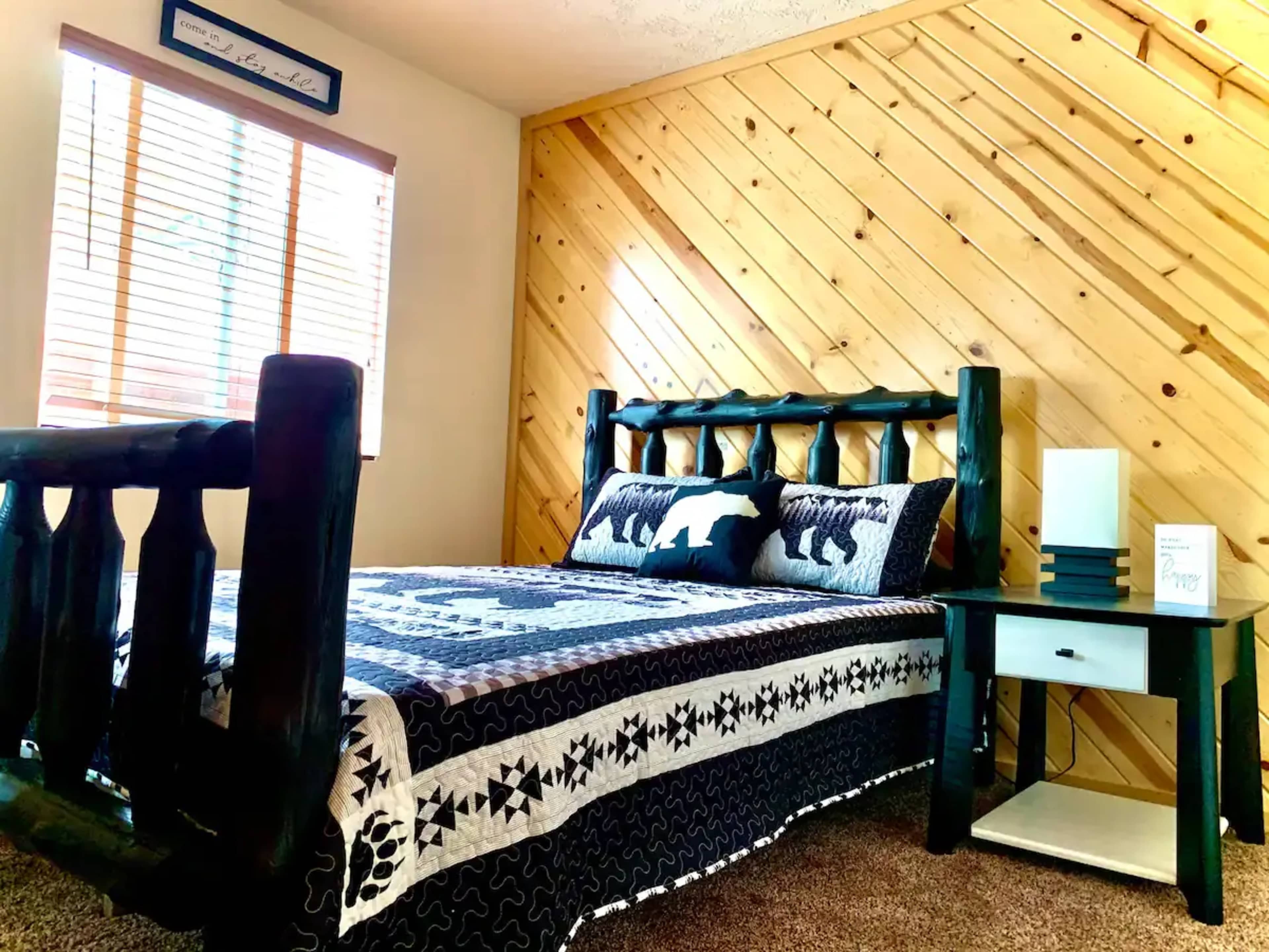 The image shows a cozy bedroom featuring a wooden bed with a black and white patterned quilt, a nightstand, and a wall with wooden paneling.