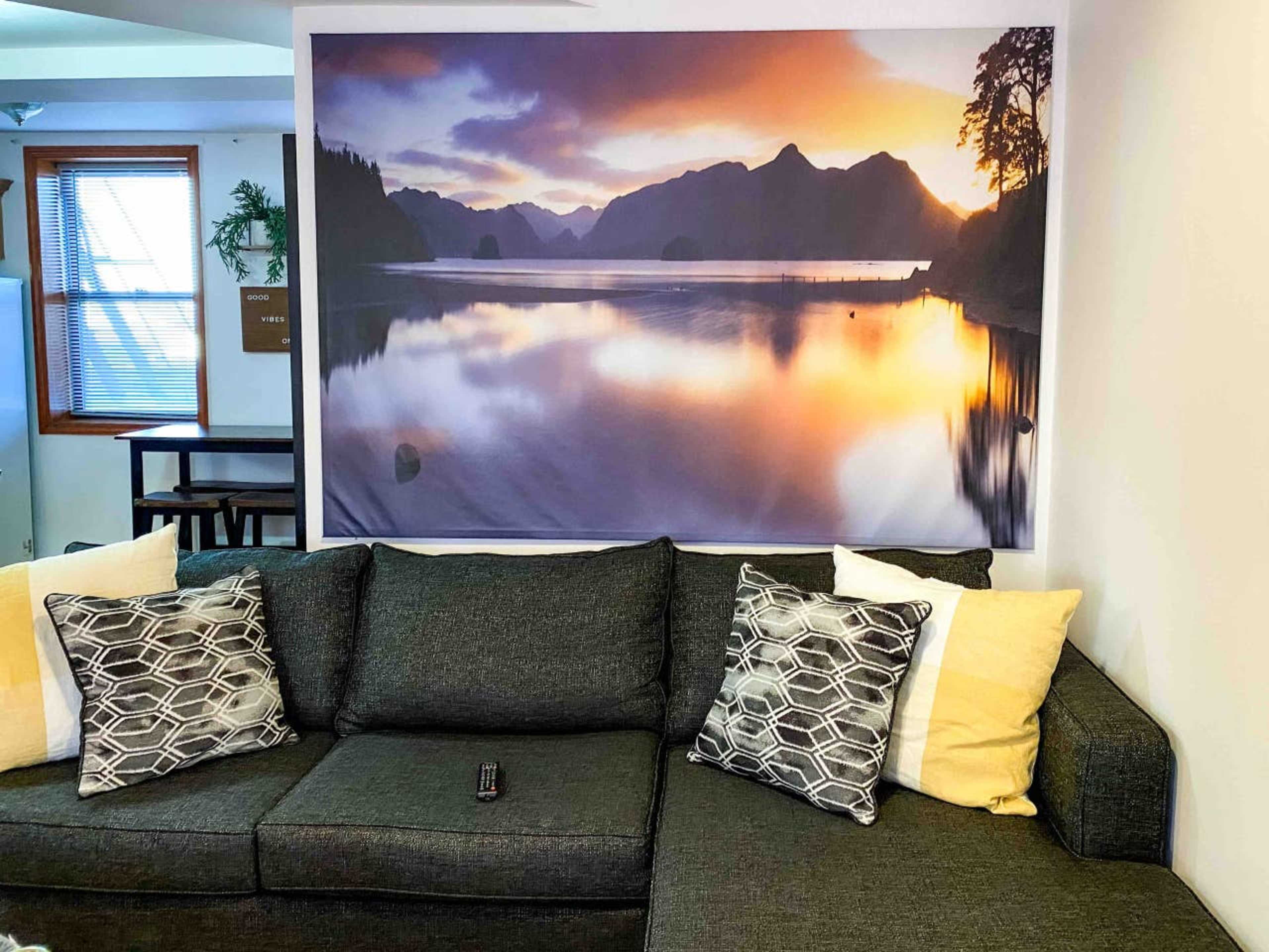 A dark gray couch with patterned cushions sits in front of a large framed landscape photograph of a serene lake surrounded by mountains at sunset.