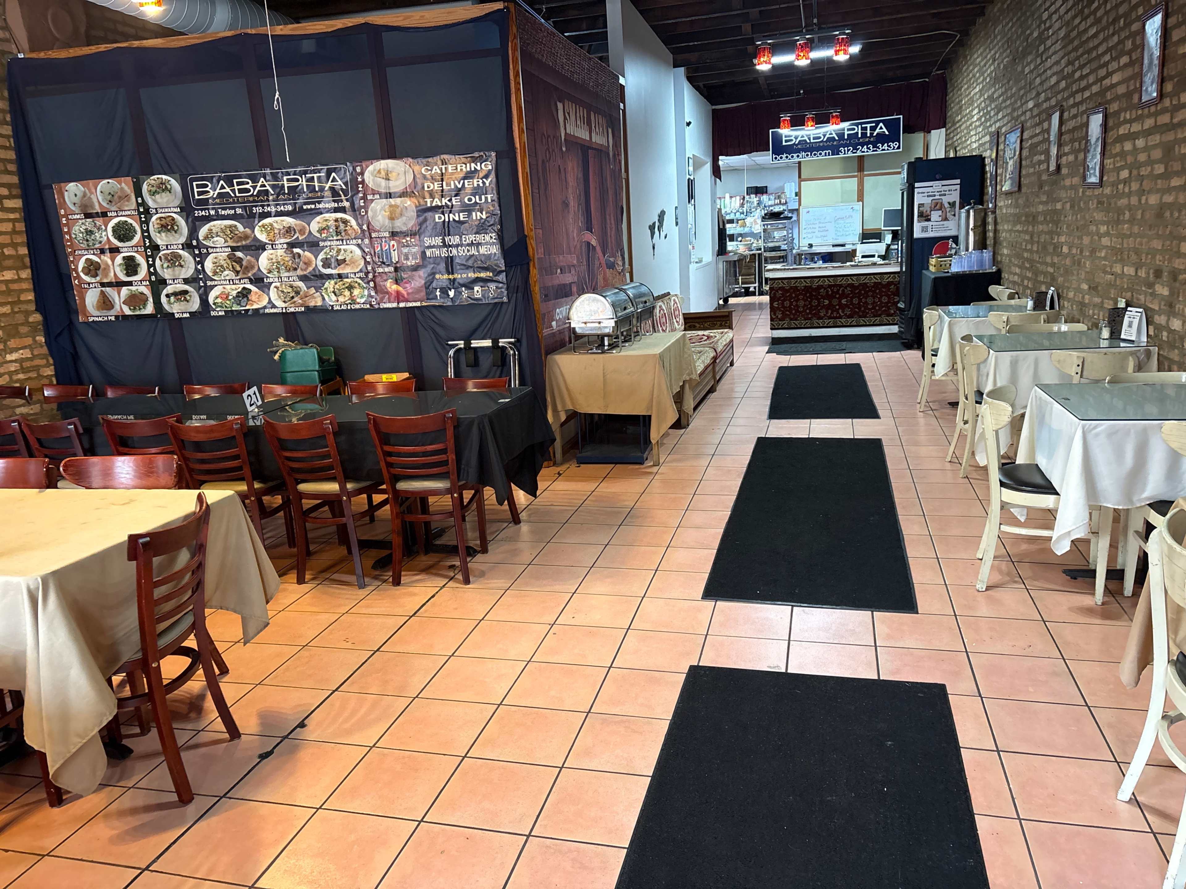 The image shows the interior of a restaurant with tables and chairs arranged on either side of a central walkway, featuring a menu display on the left wall and a counter at the end.
