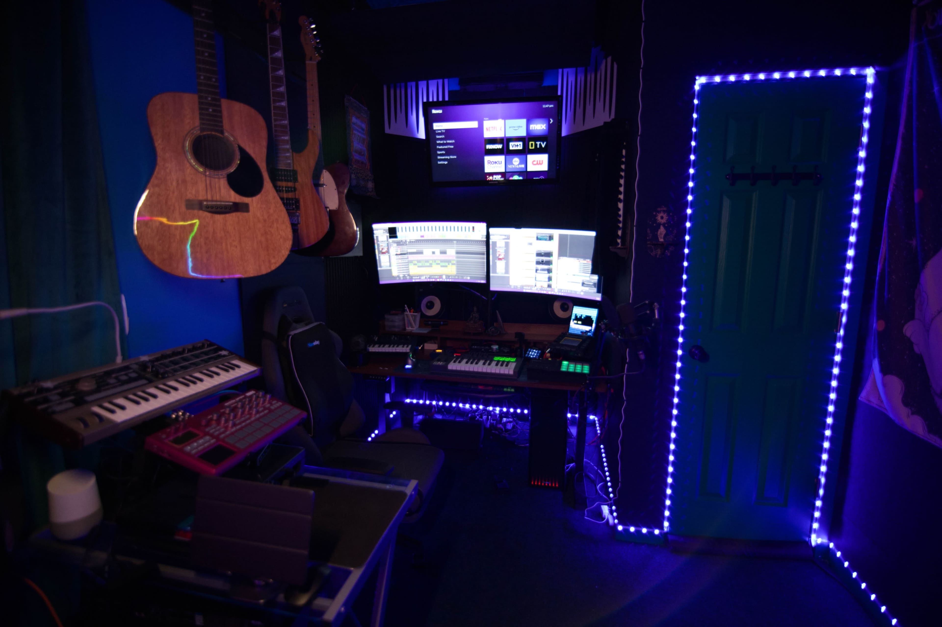 The image shows a music production room with various instruments, including guitars and keyboards, alongside dual computer monitors and colorful LED lighting.
