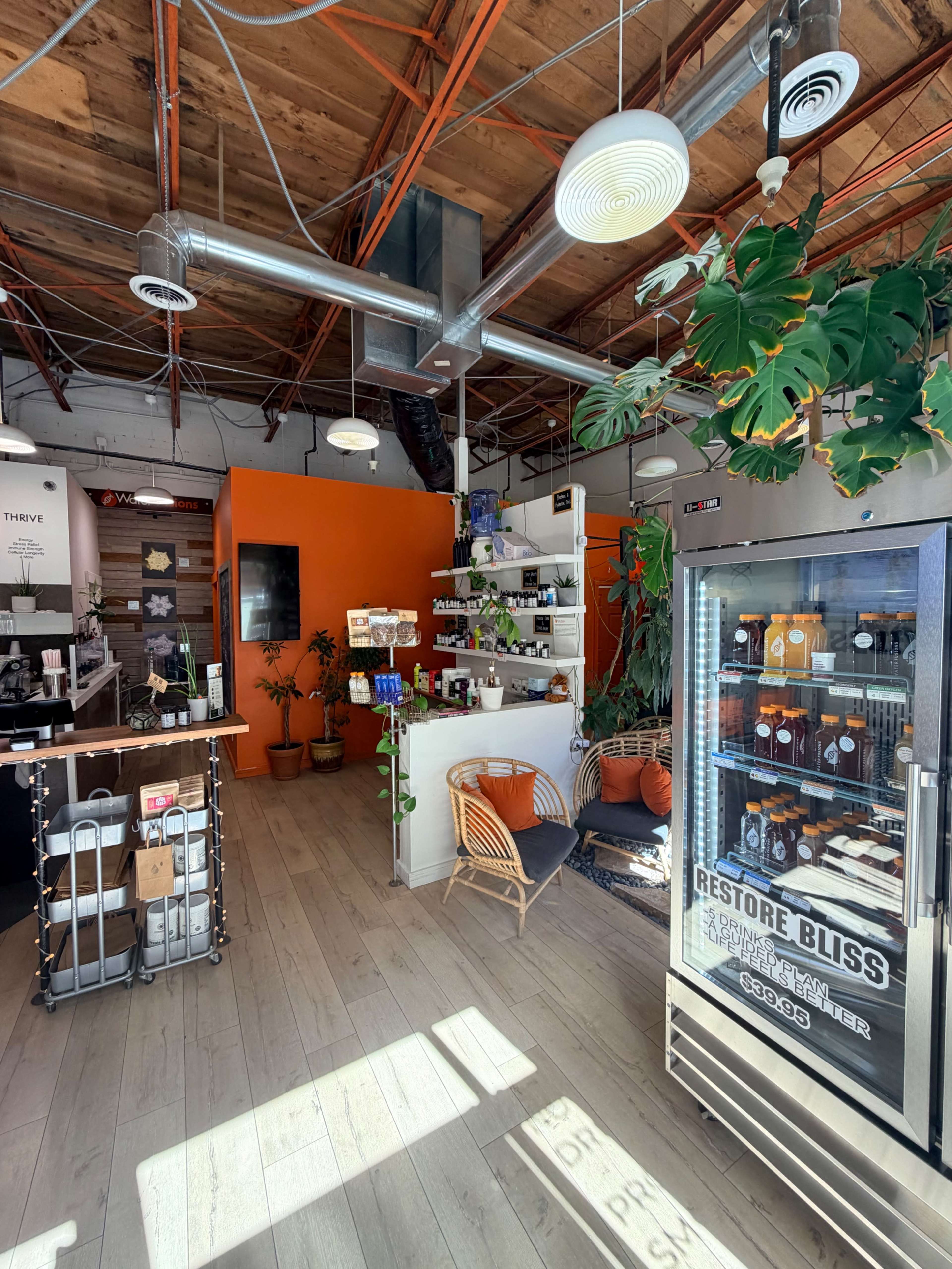 The image shows a bright, open space of a modern café or wellness shop with orange accents, shelves filled with products, and a display fridge on one side.