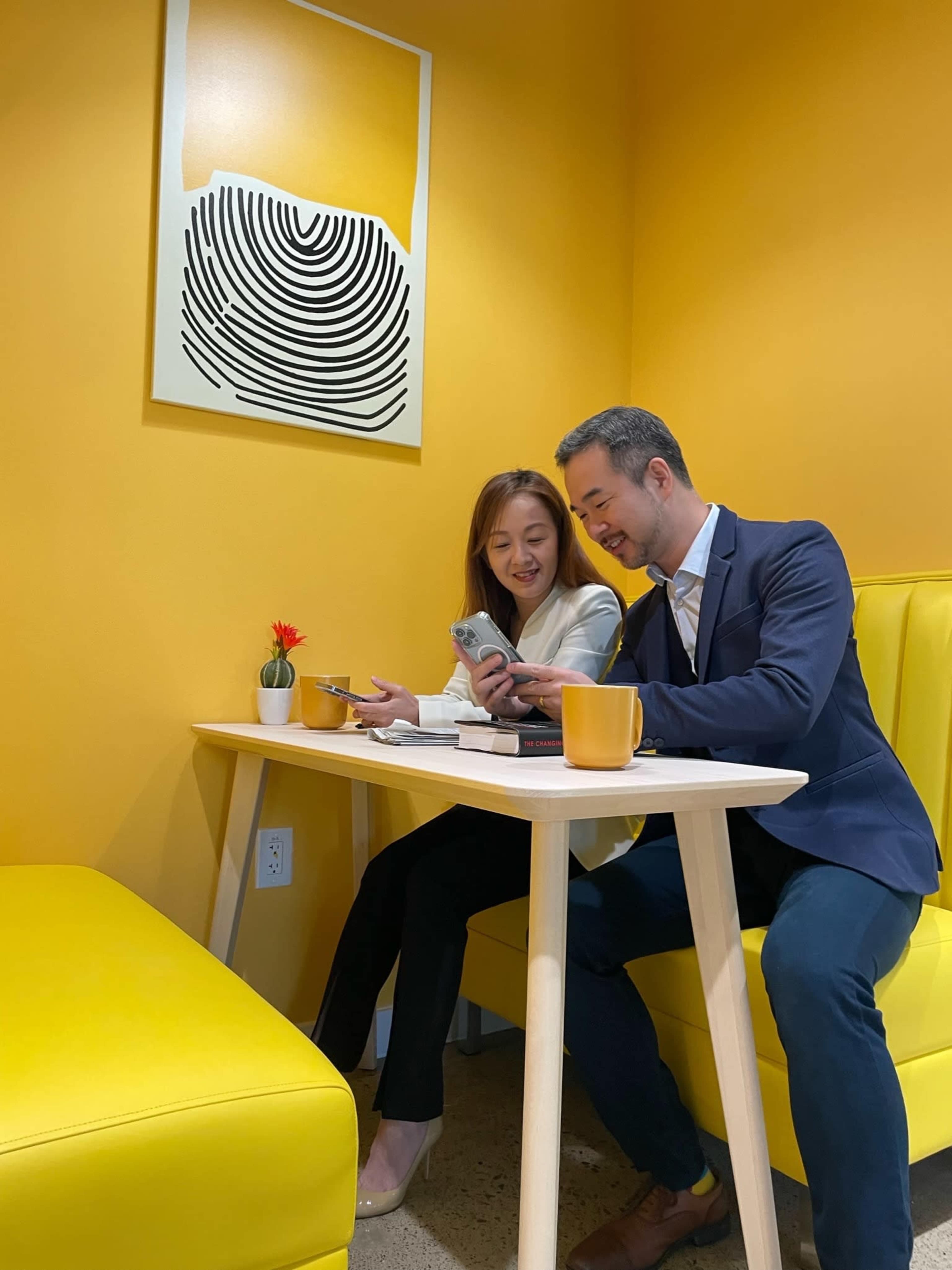 A man and a woman are seated at a small table in a bright yellow room, looking at a smartphone together.