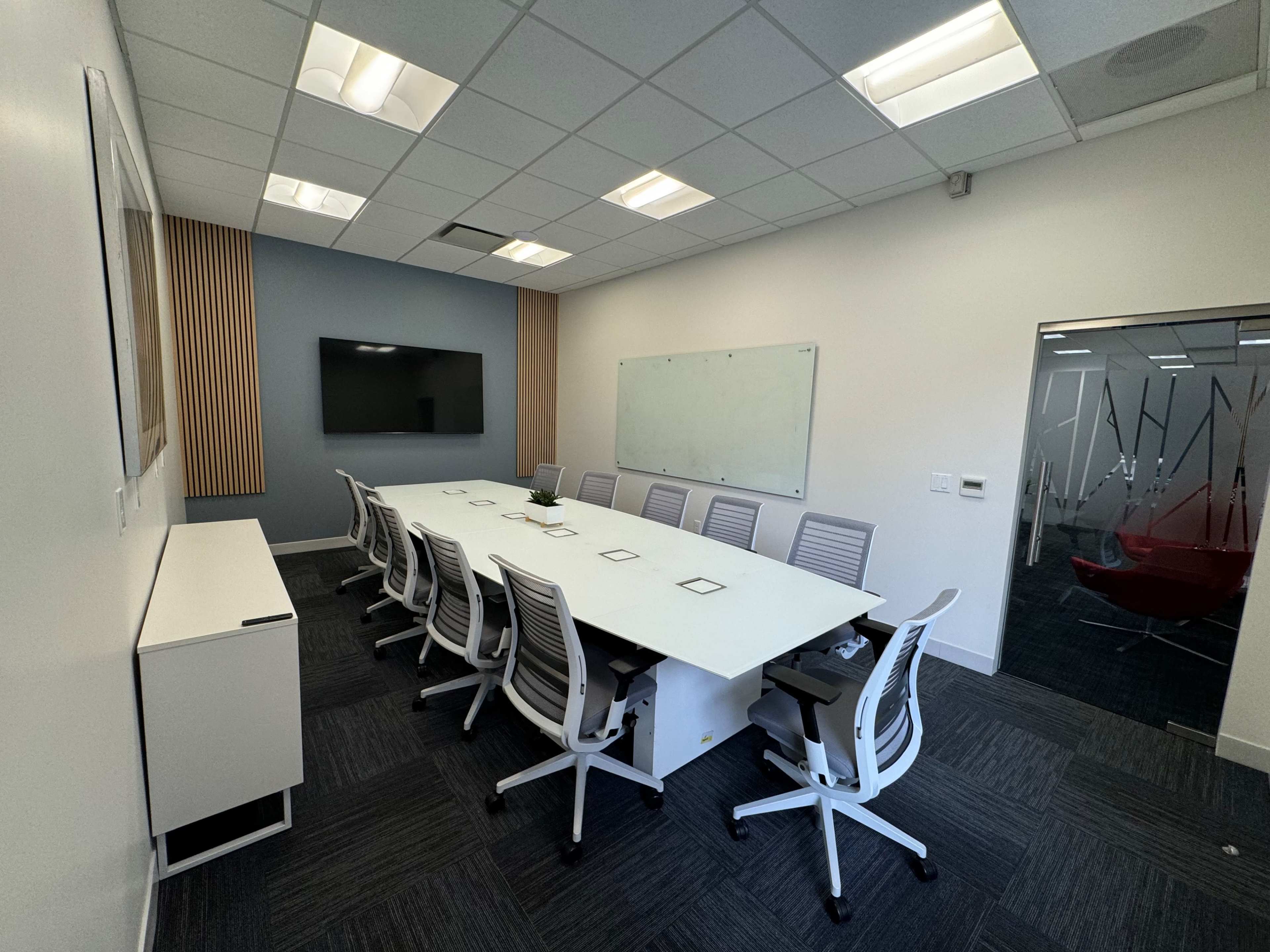 A modern conference room featuring a long white table surrounded by ergonomic chairs, a wall-mounted television, and a whiteboard.