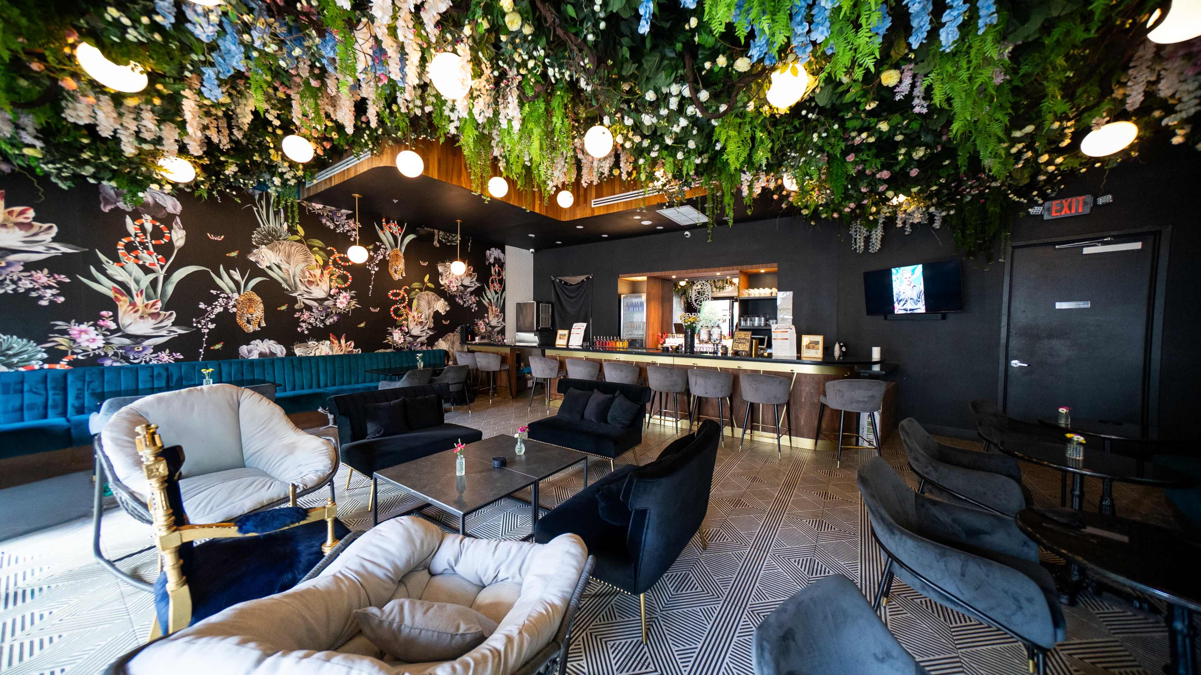 The image shows a stylish café interior with floral wallpaper, hanging decorations, a well-lit bar area, and various seating arrangements including plush chairs and tables.