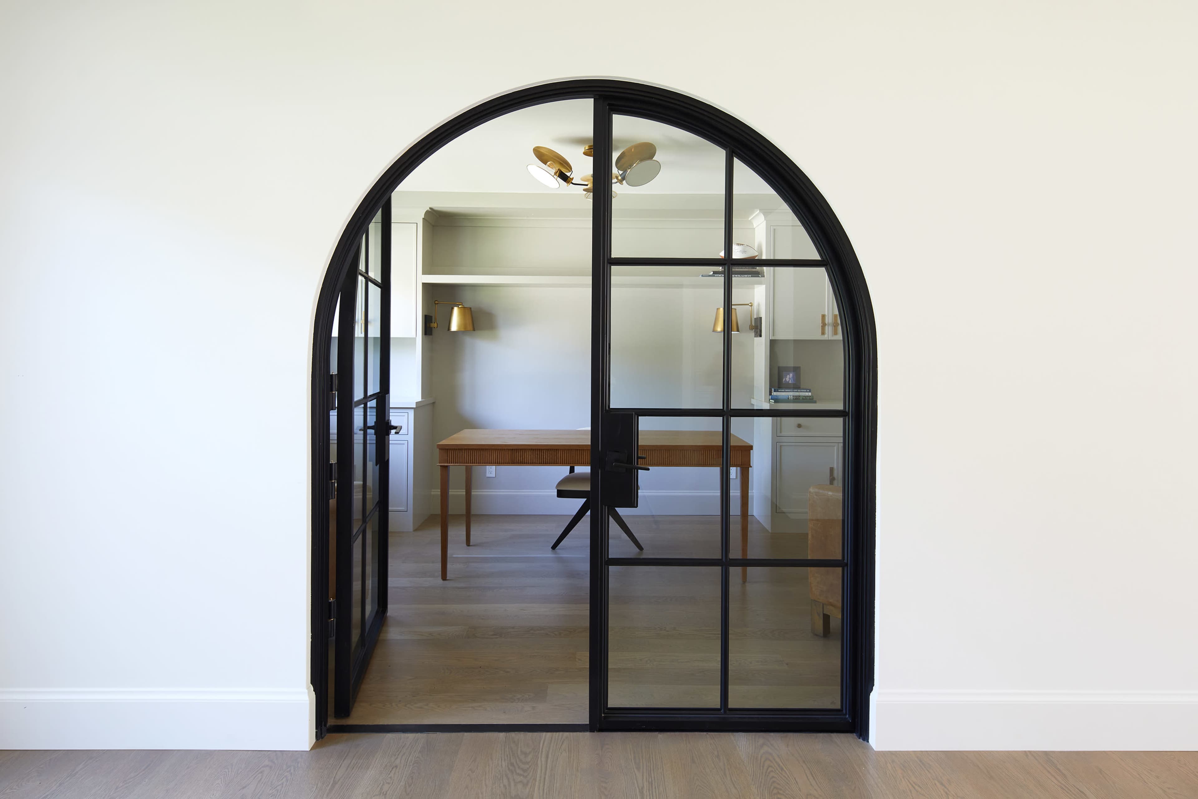 A curved, black-framed glass door leads into a room featuring a wooden desk and wall shelves.