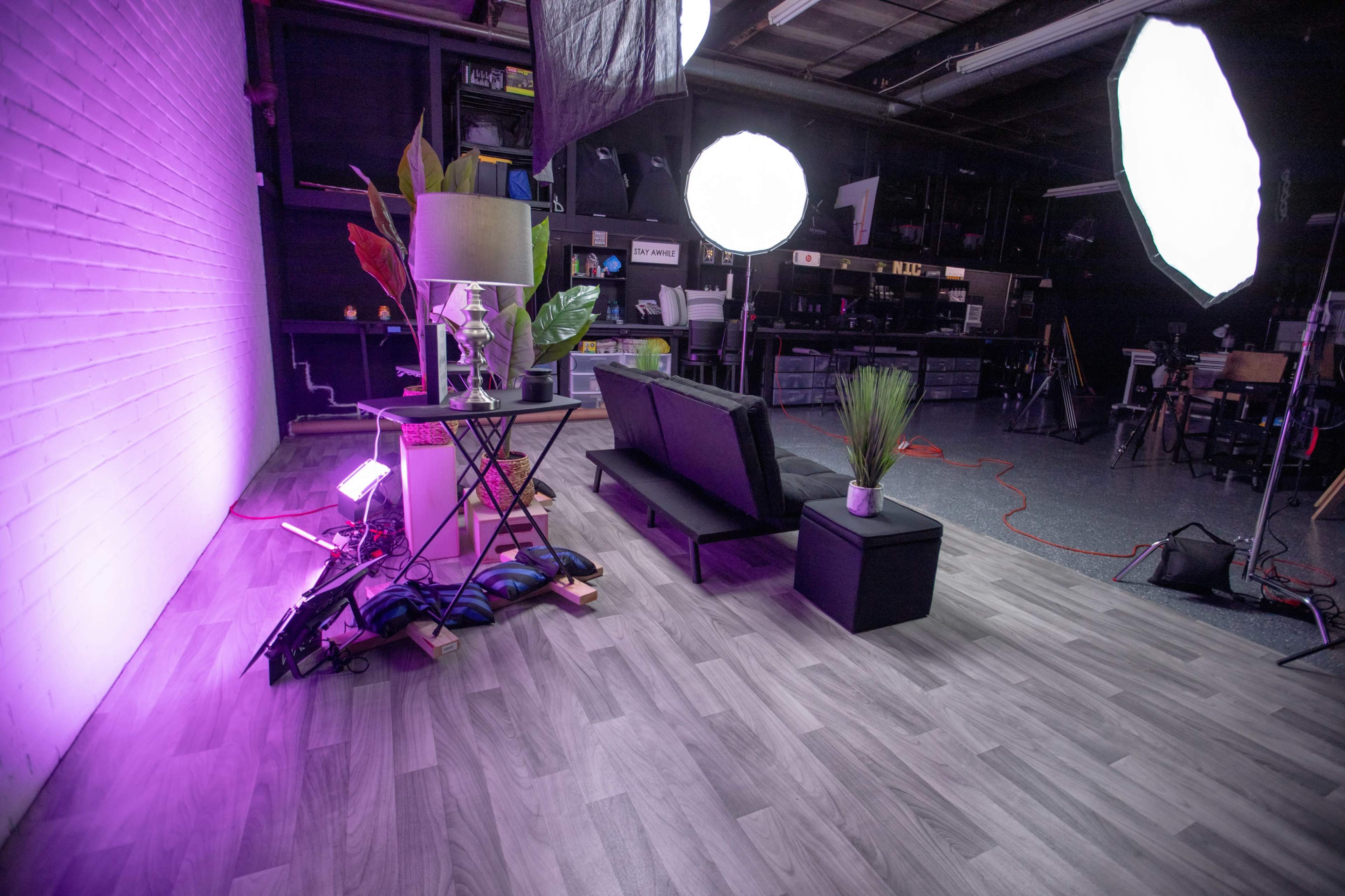 A modern studio setup featuring a black couch, a table with a lamp and potted plants, and large lighting equipment against a white-brick wall.
