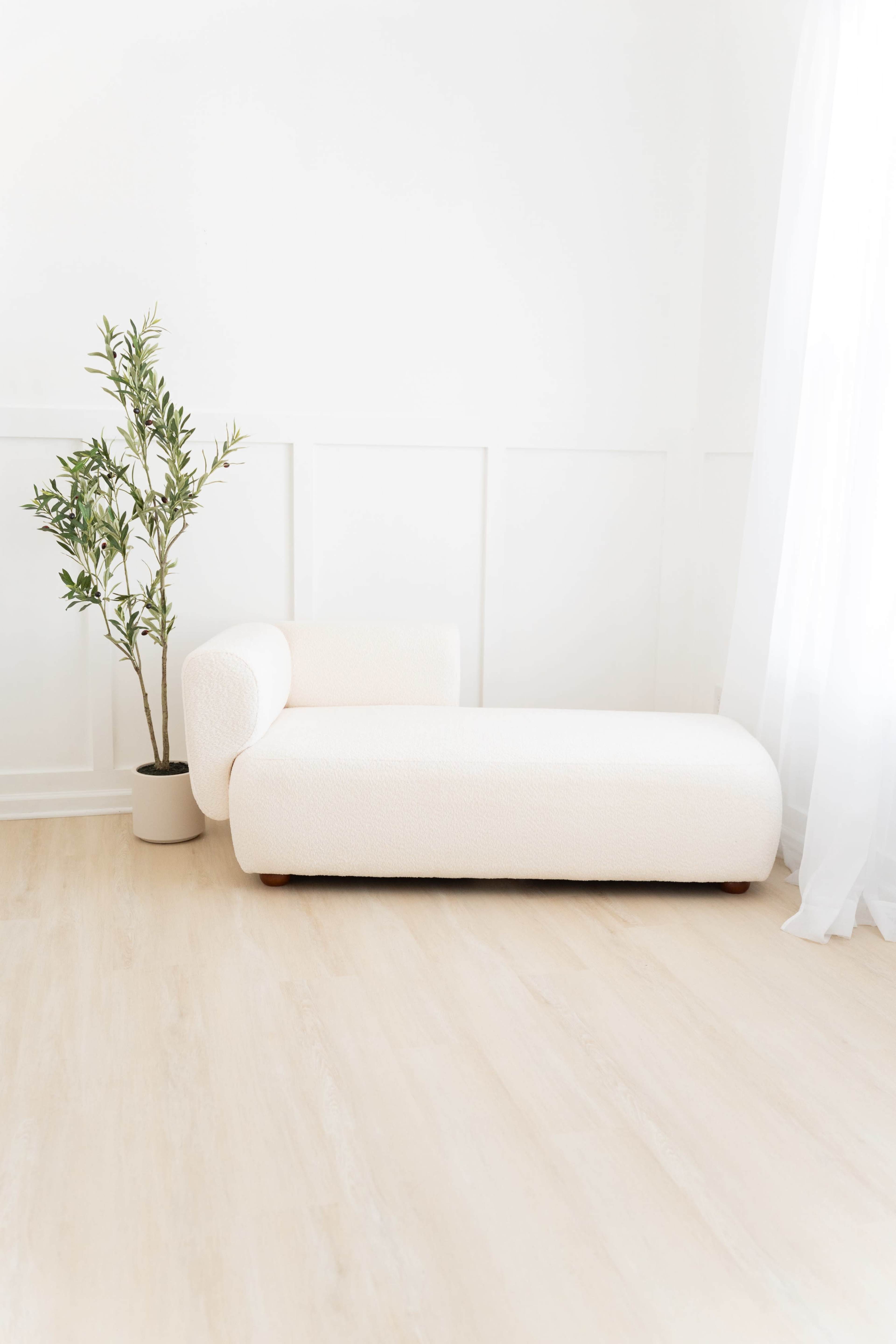 A light-colored chaise lounge is positioned against a white wall, next to a potted plant.