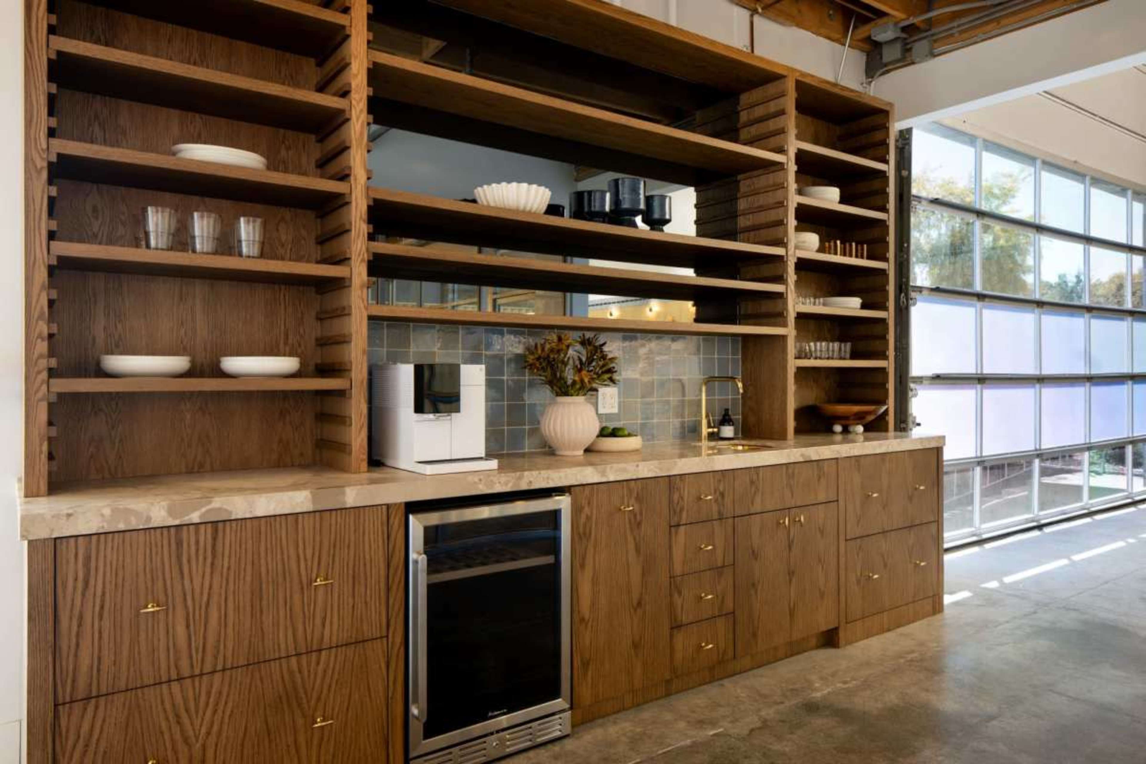 A modern kitchen features wooden open shelves, cabinets, a coffee maker, and a wine cooler, with a large window allowing natural light.