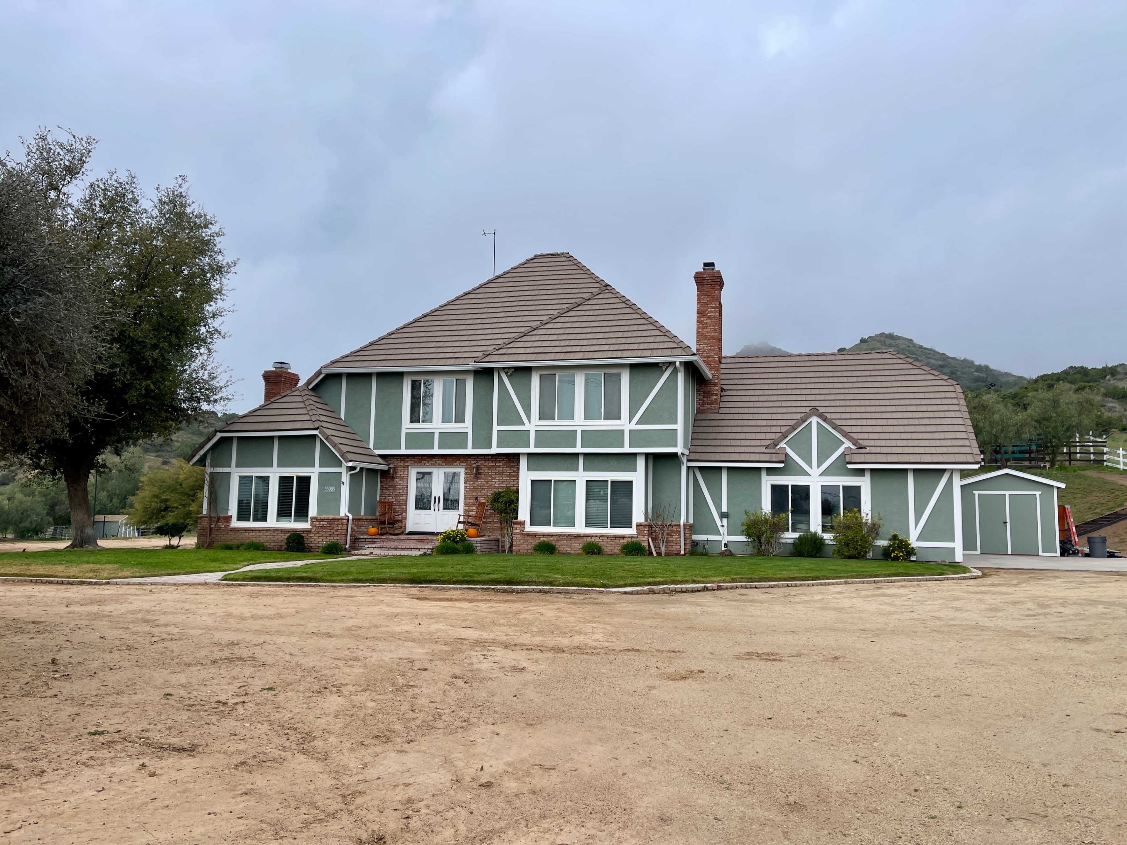A large, two-story green house with a brown tiled roof and brick accents is situated on a dirt lot surrounded by trees and hills.