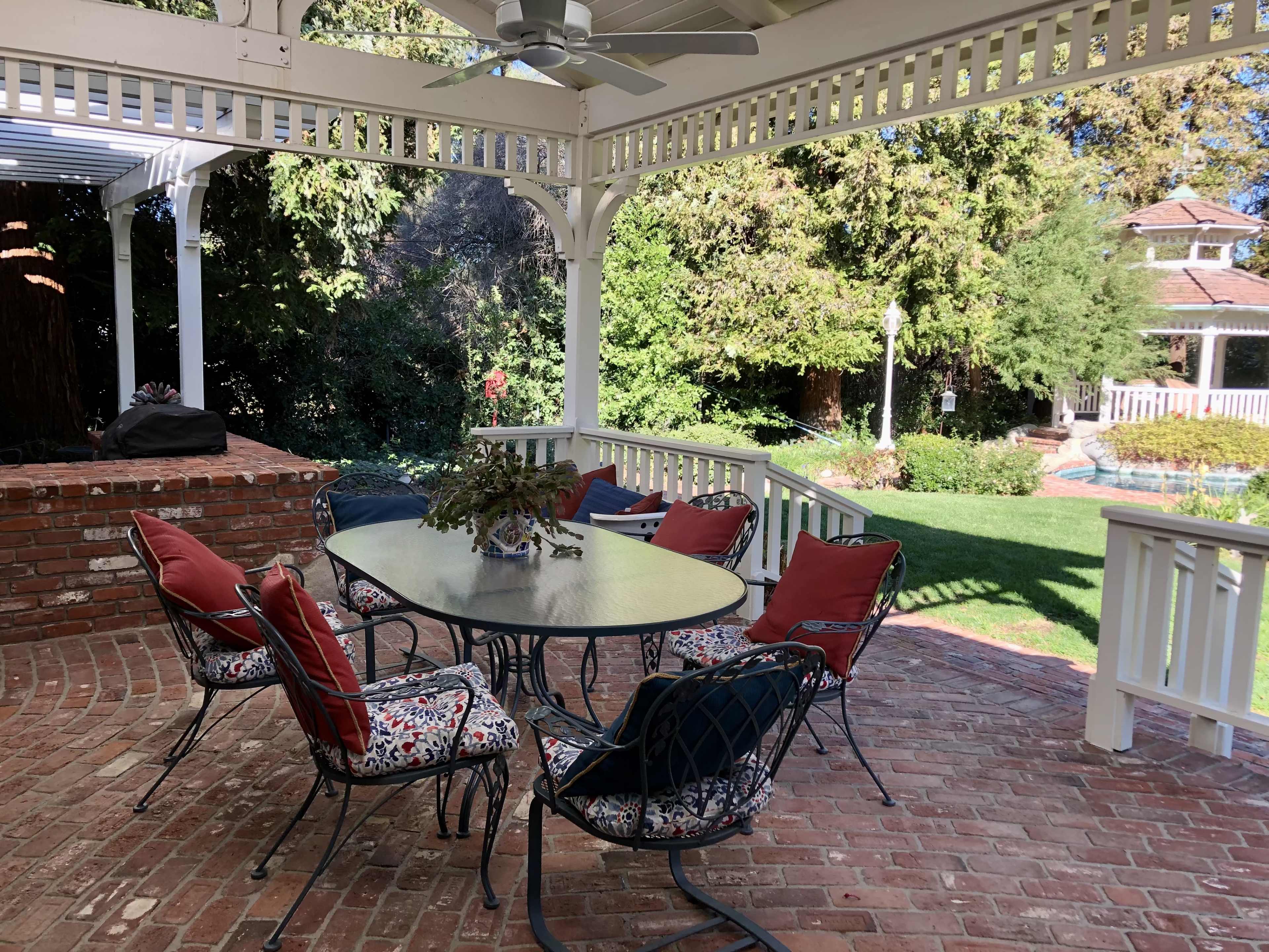 A patio dining area features a round table surrounded by six chairs with colorful cushions under a white pergola.