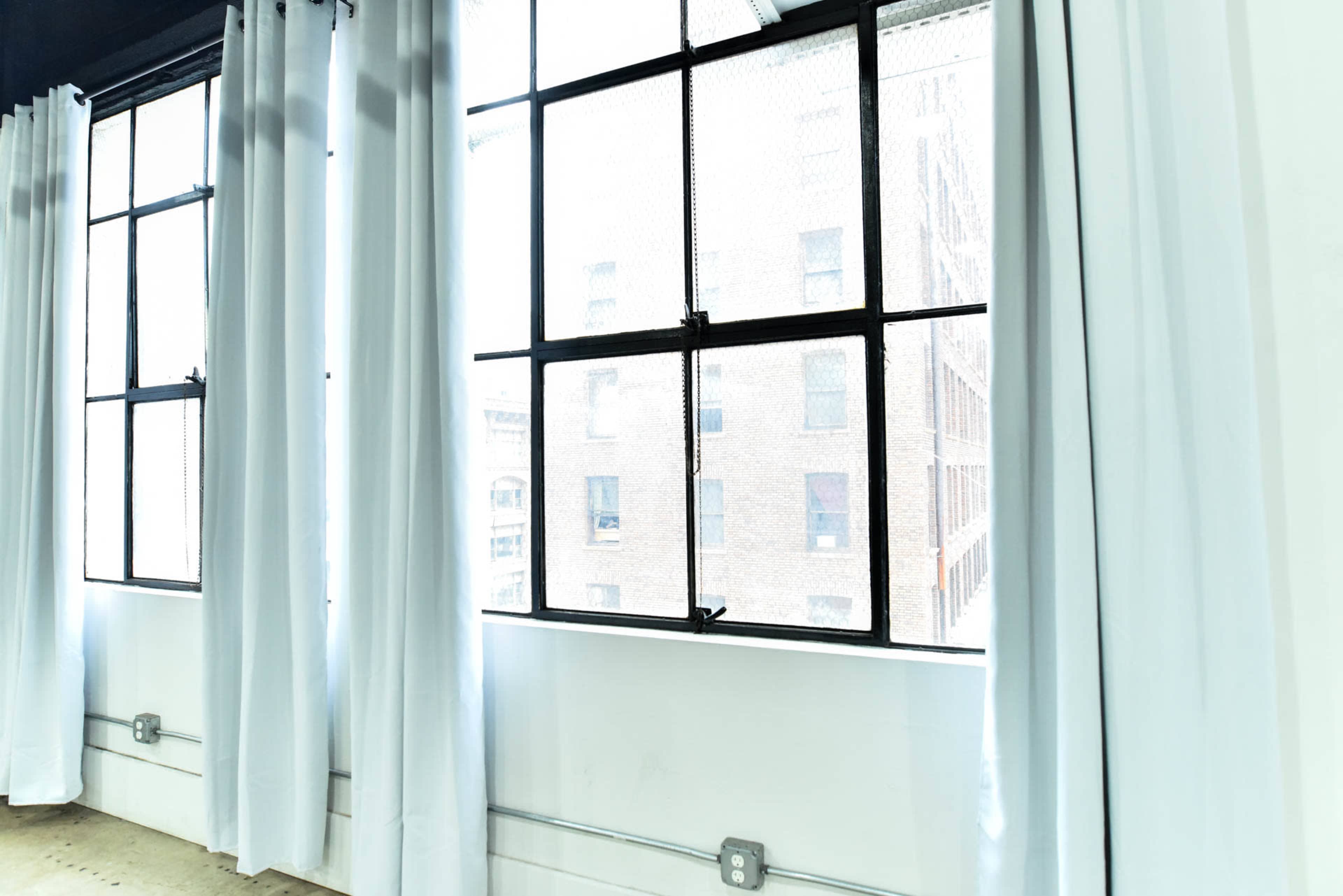 The image shows a bright room with large windows framed by white curtains, revealing a view of a brick building outside.