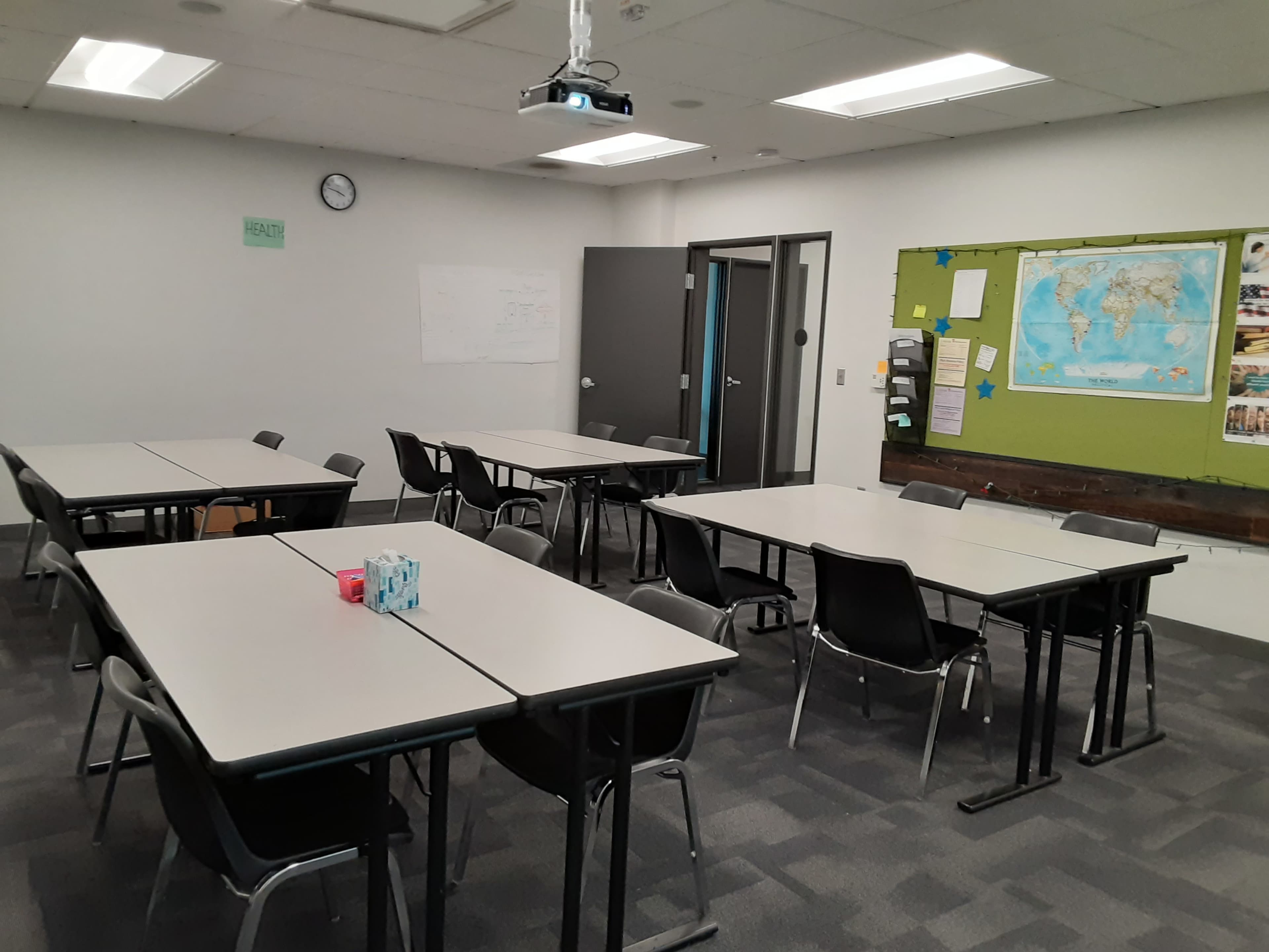 A classroom setup features several tables and chairs arranged for collaborative work, with a projector and maps visible on the walls.