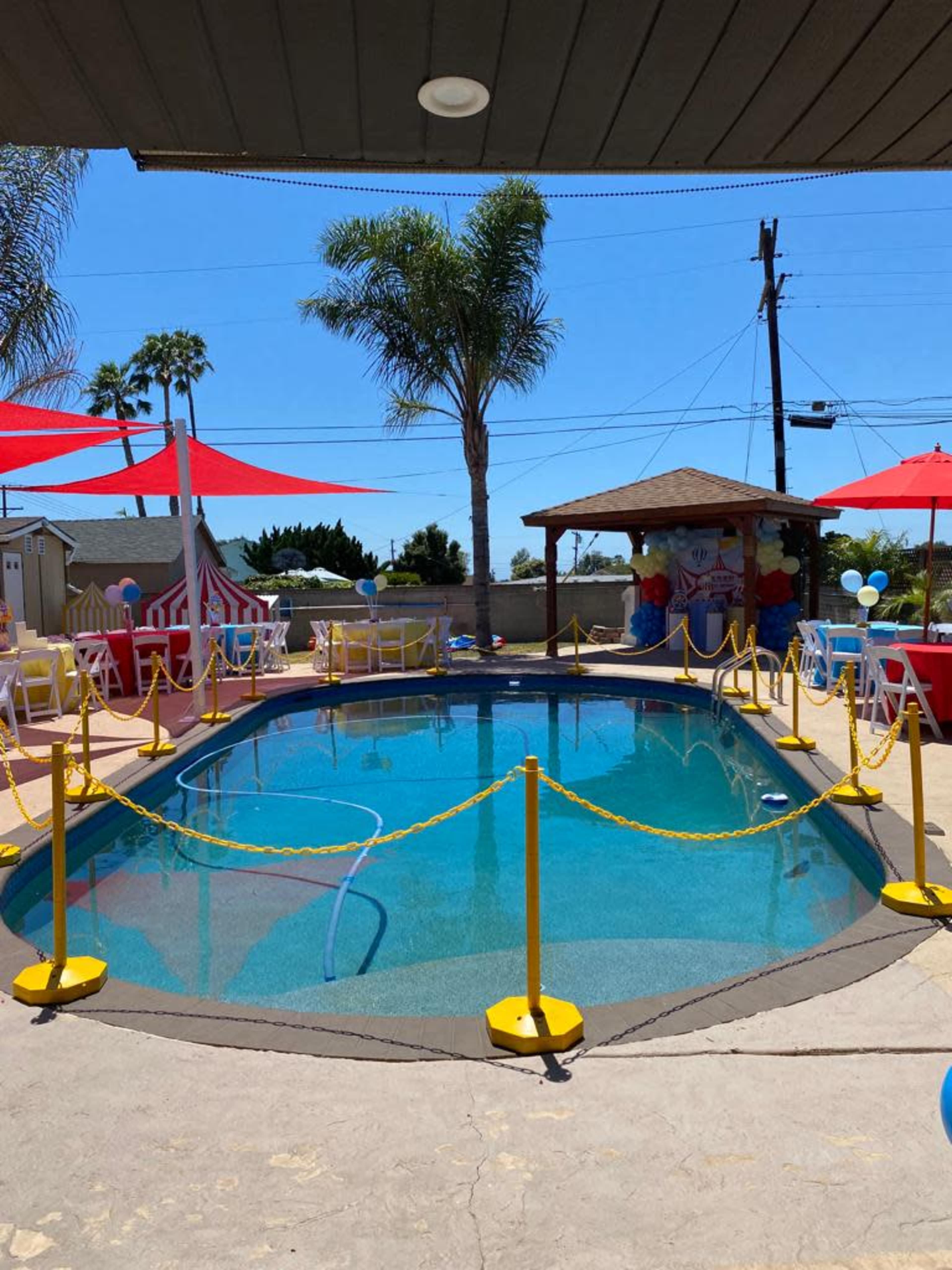 The image shows a swimming pool surrounded by colorful chairs, canopies, and a gazebo, set in a sunny outdoor environment.