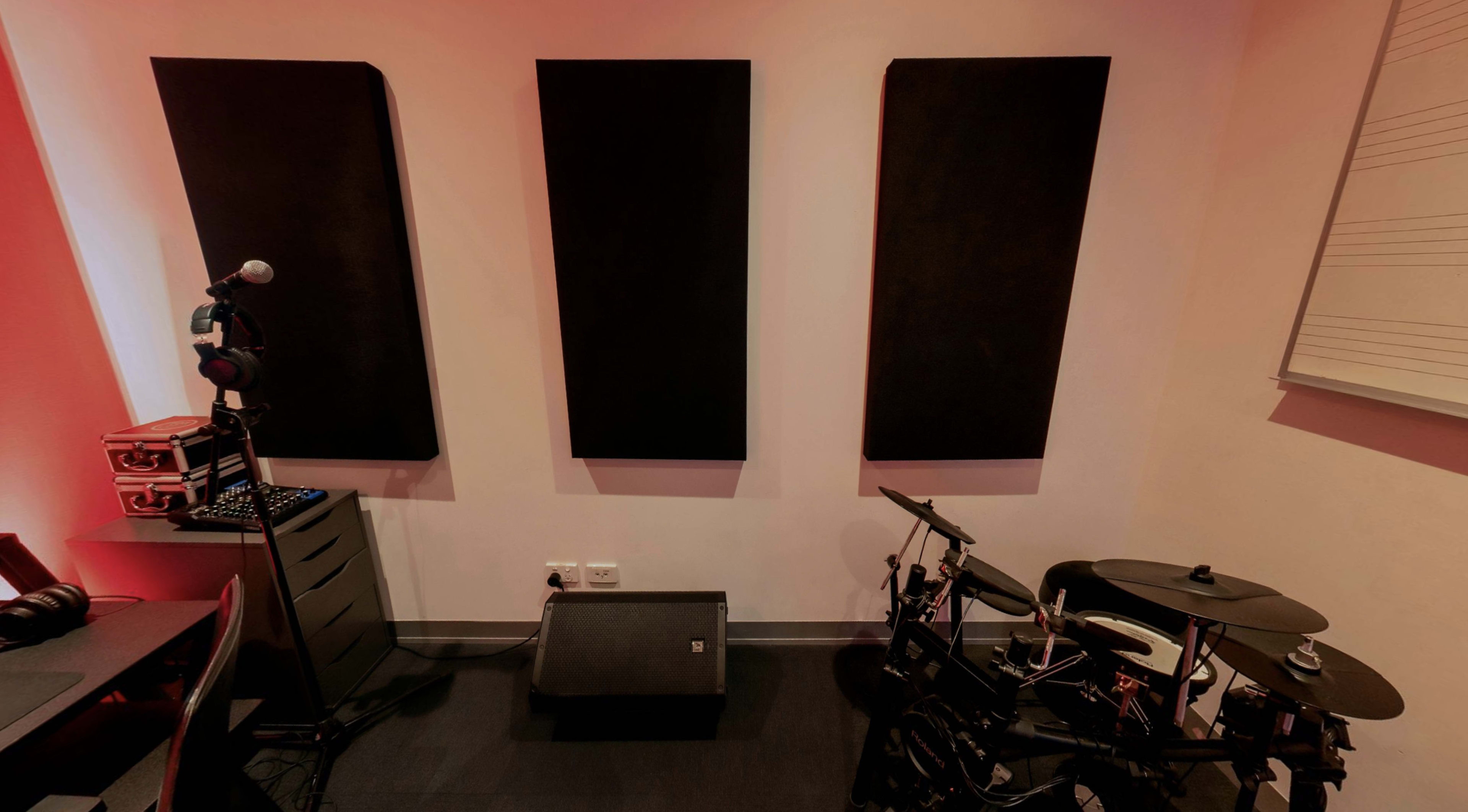 A music studio corner with three black acoustic panels on the wall, a microphone, an electronic drum set, and a speaker positioned on the floor.