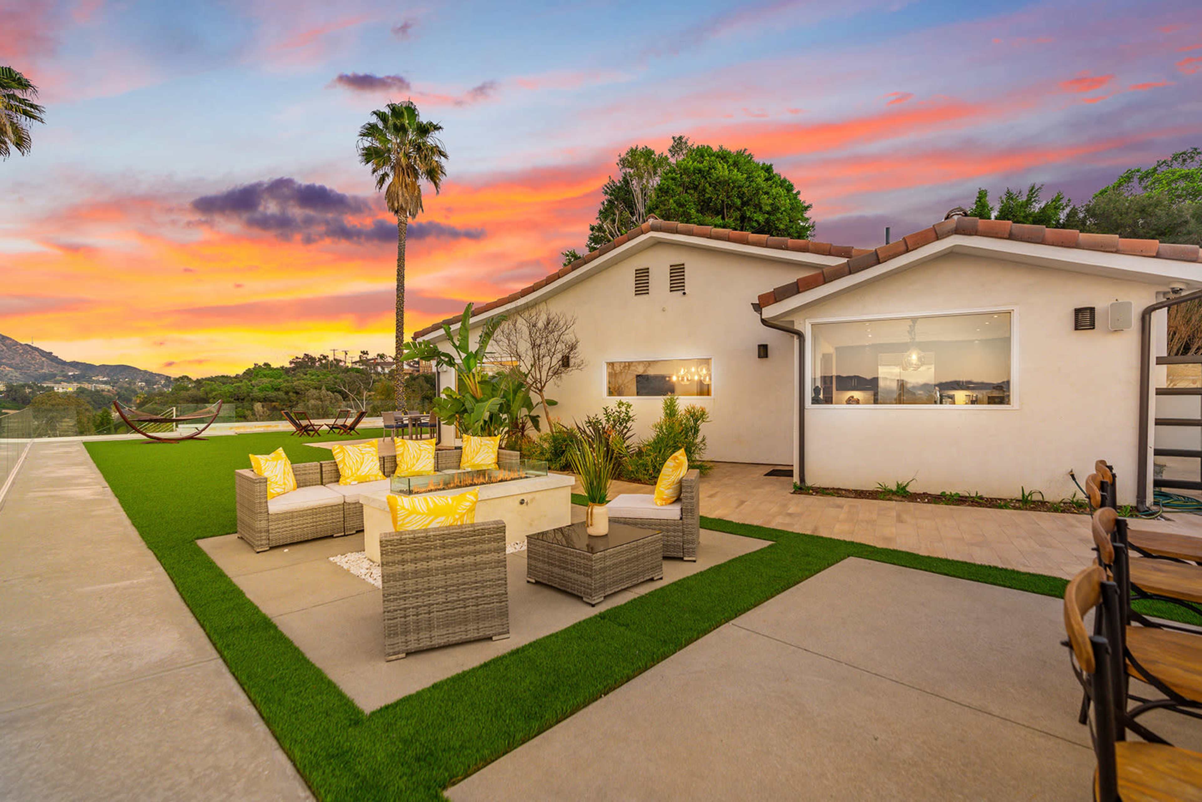A modern outdoor seating area is set on a green lawn beside a house, with palm trees and a colorful sunset in the background.
