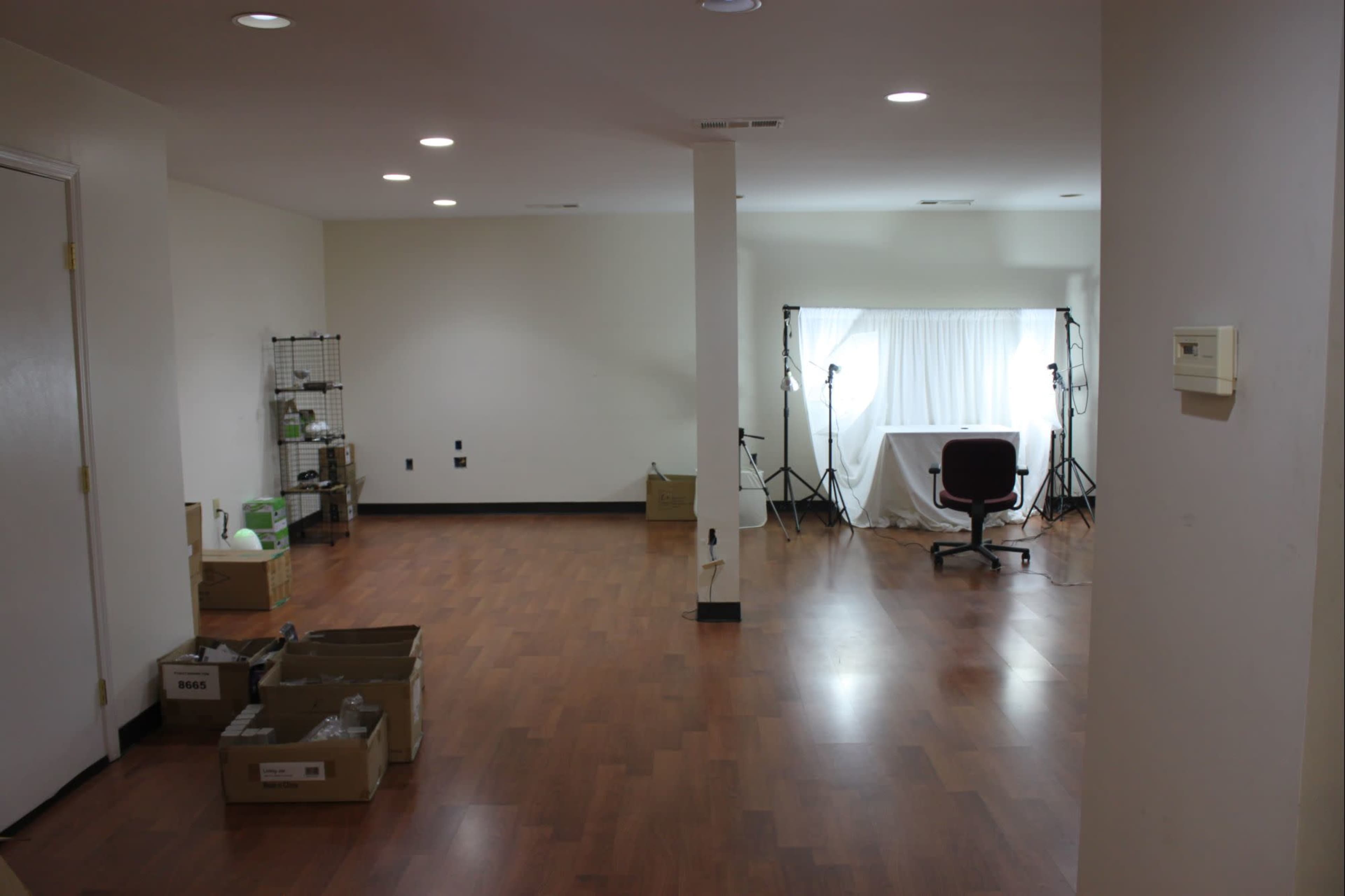 The image shows a spacious, empty room with wood flooring, a window covered by light curtains, and a chair positioned near photography equipment on stands.