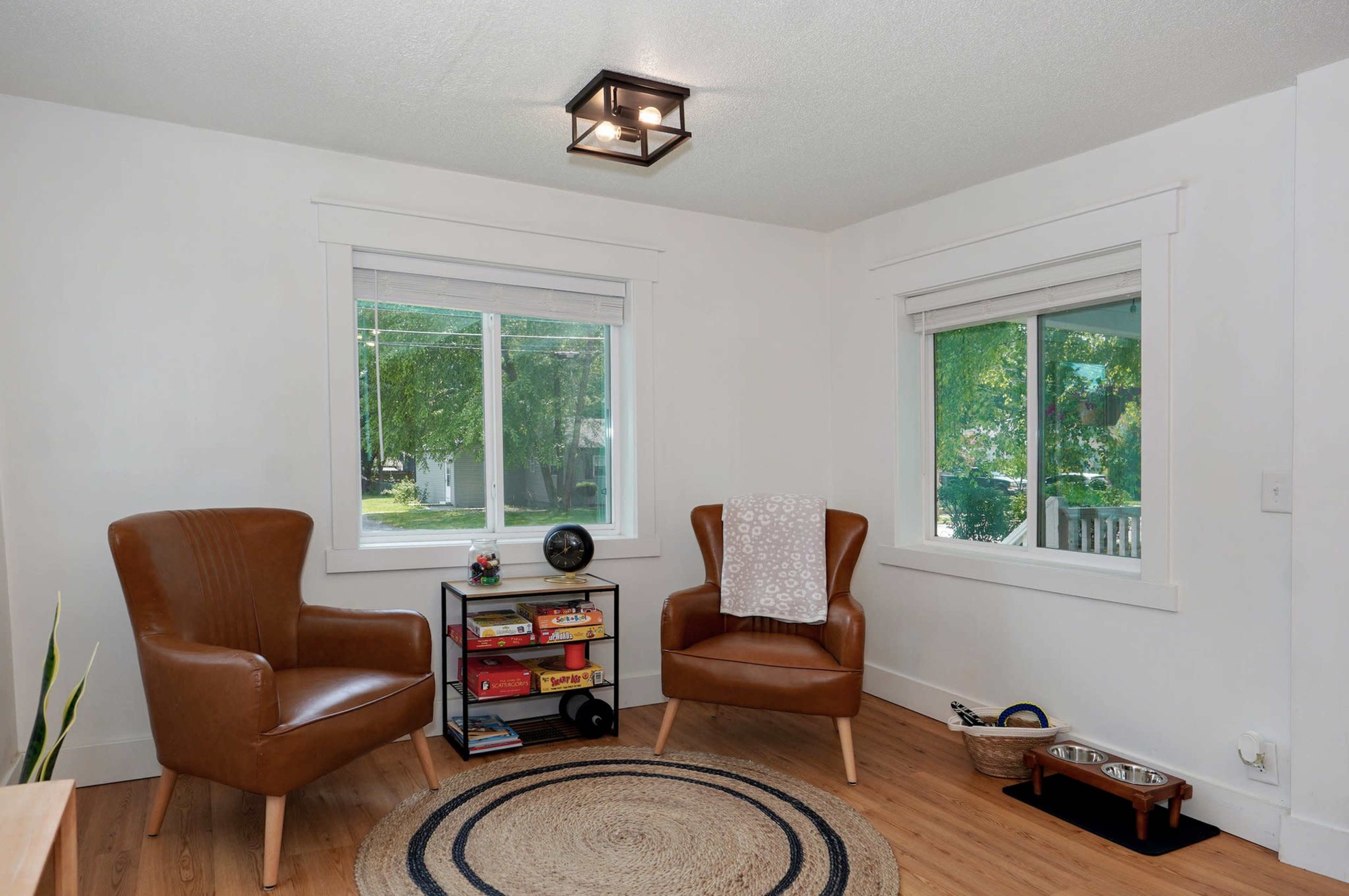 The image shows a bright, minimalist room featuring two brown armchairs, a small bookshelf, a circular rug, and two windows that provide natural light.