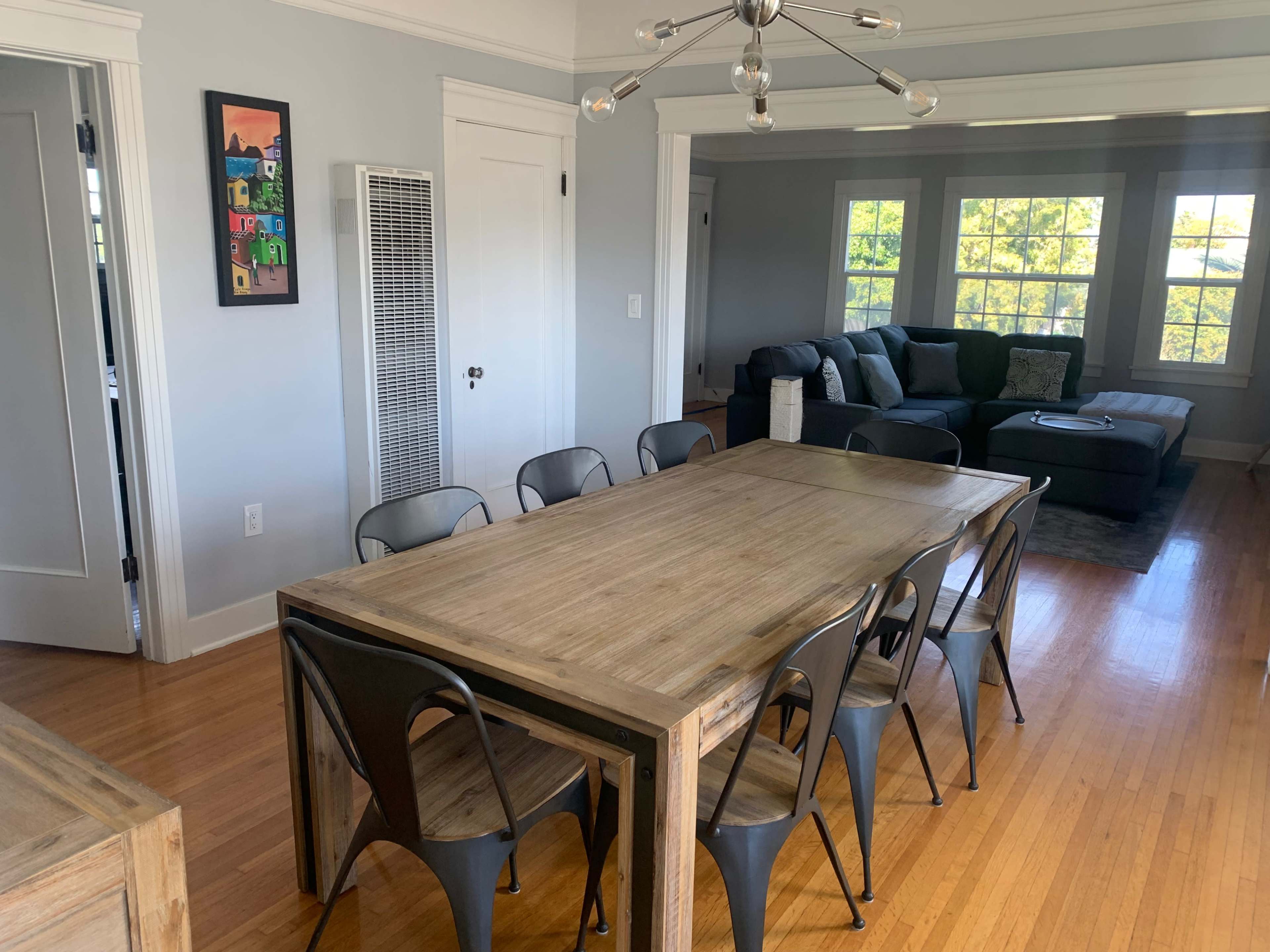 A wooden dining table with metal chairs is positioned in a brightly lit dining area adjacent to a living room featuring a dark sofa and large windows.