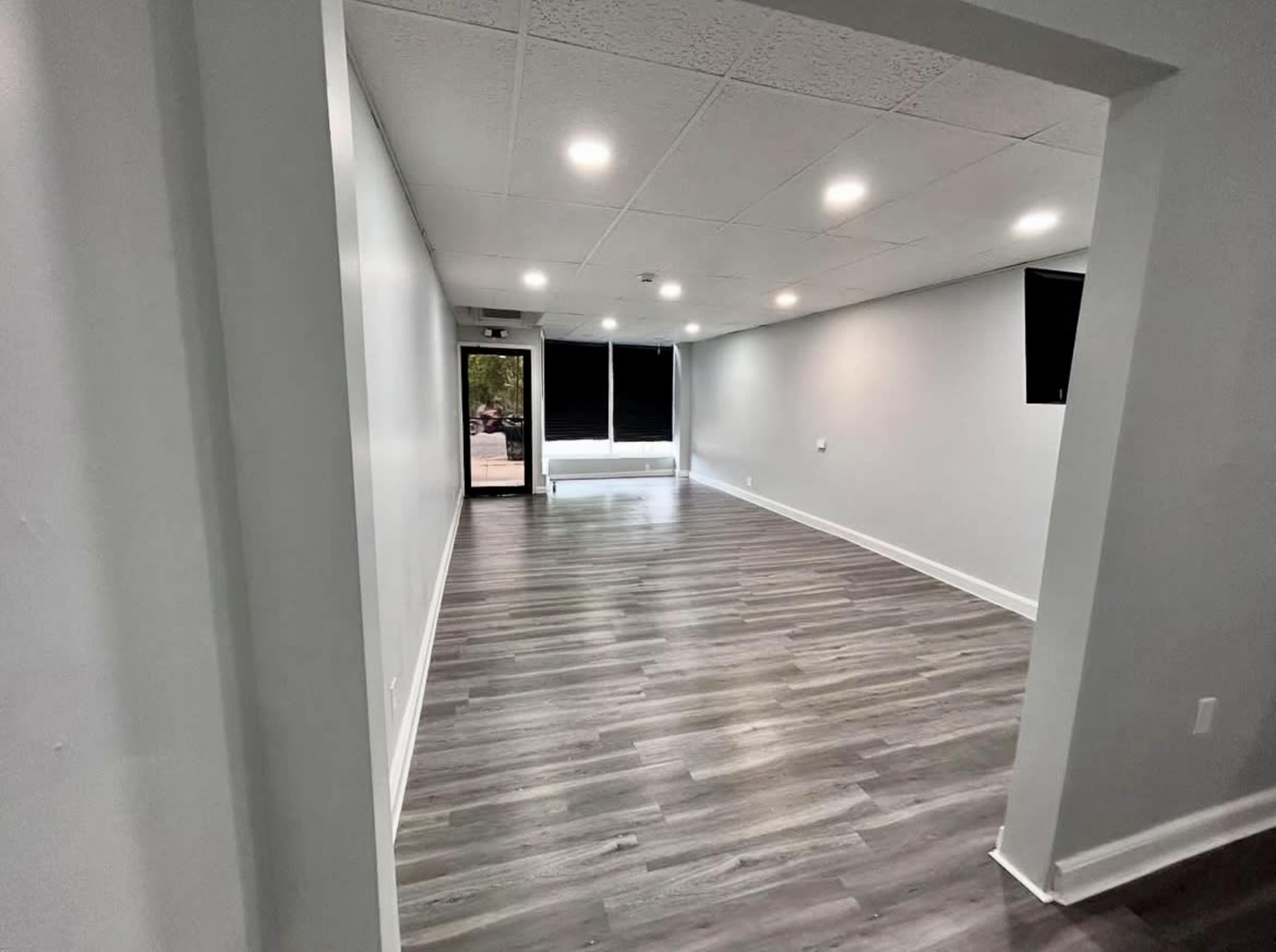 The image shows a spacious, empty room with gray walls, wooden flooring, and several overhead lights.
