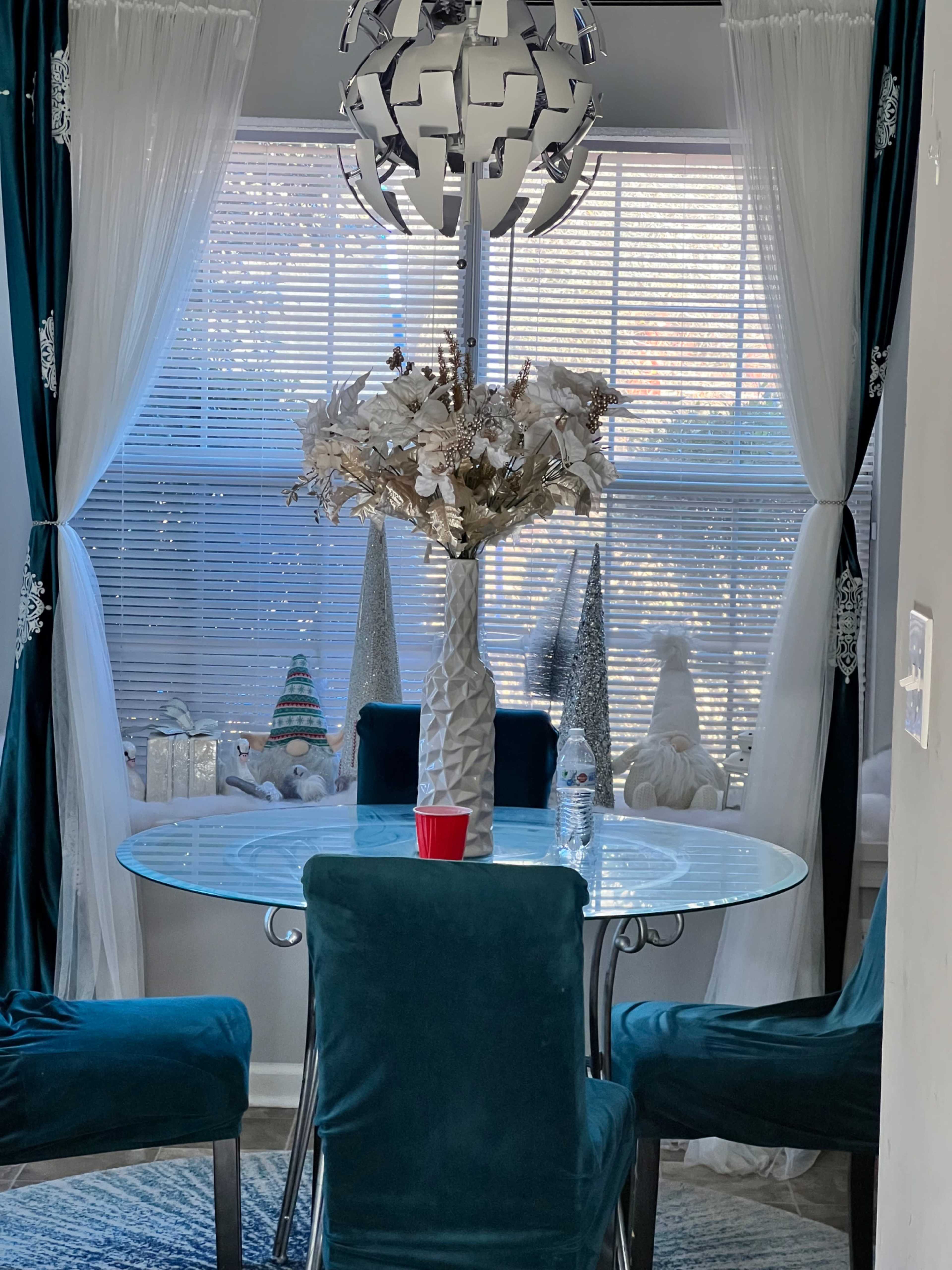A round glass table with teal velvet chairs is set in a bright dining area decorated with a white floral arrangement and holiday-themed accents.