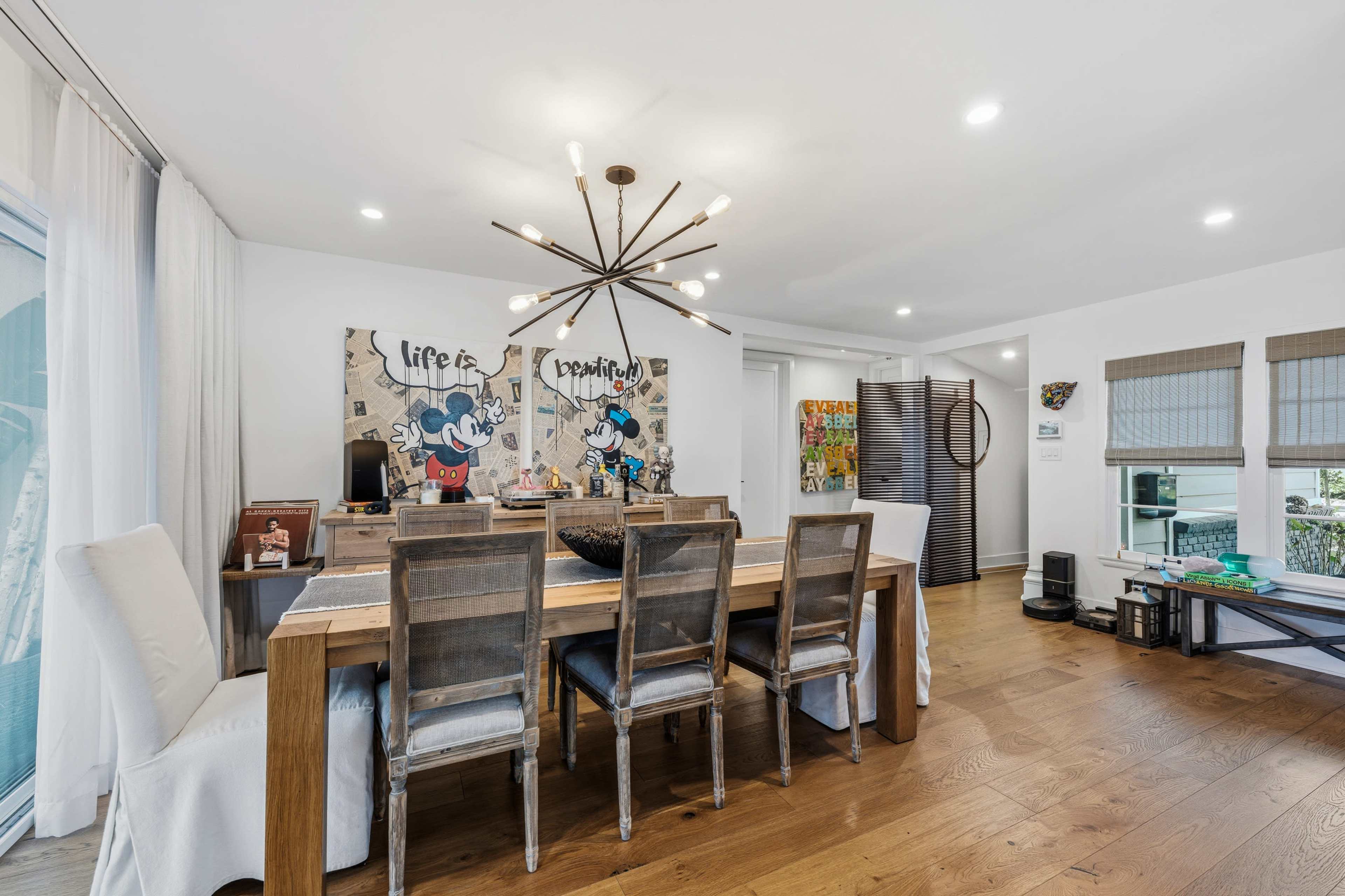 The image shows a modern dining room with a large wooden table, six chairs, and a contemporary light fixture, decorated with vibrant artwork on the walls.