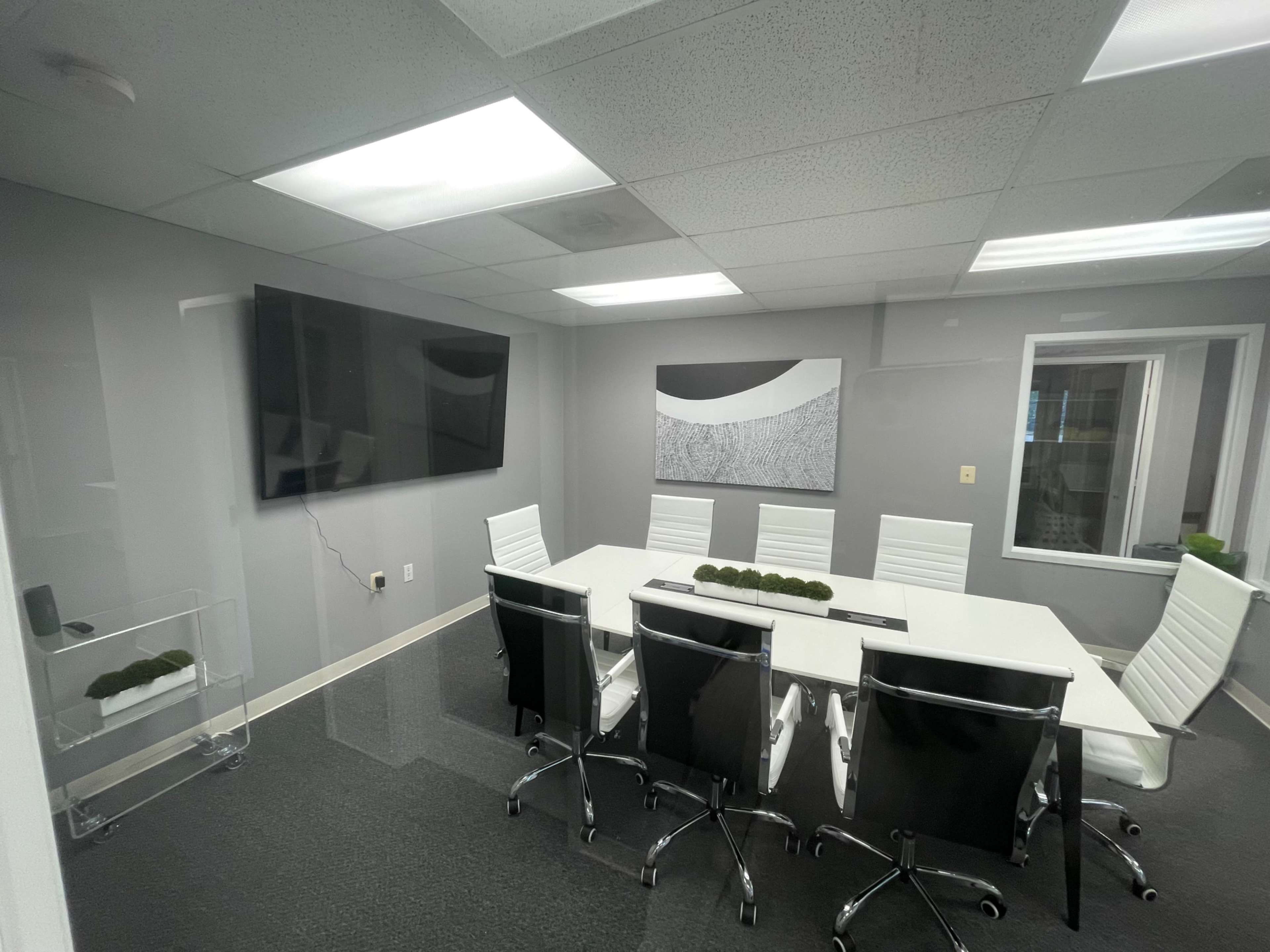 A modern conference room with a rectangular table surrounded by eight chairs, a large TV mounted on the wall, and a decorative wall piece above the table.