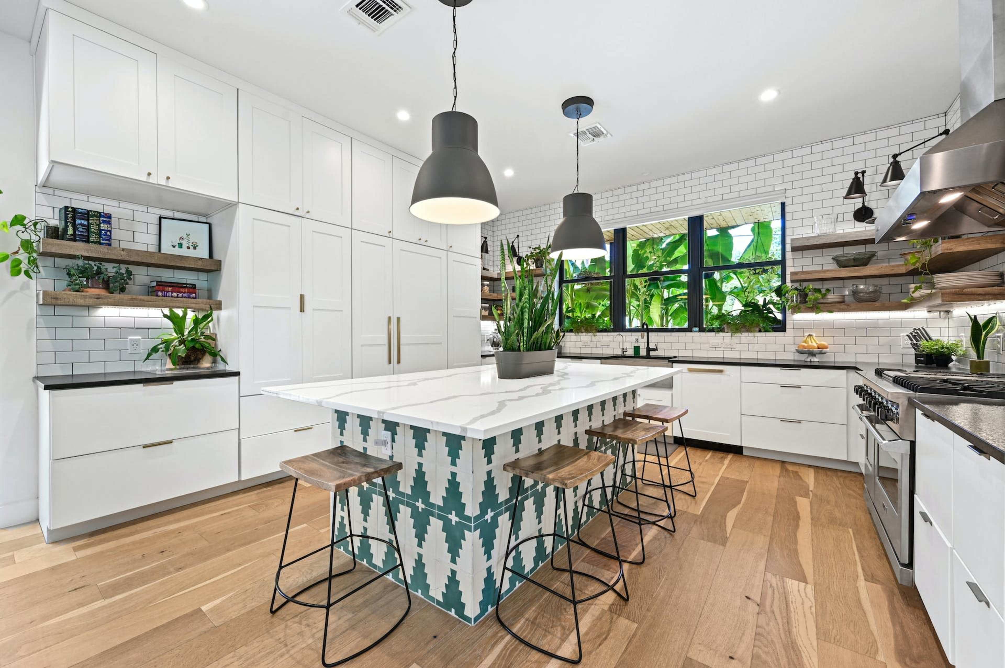A modern kitchen features a central island with a patterned tile base, surrounded by stools, and is equipped with white cabinetry and open shelving adorned with plants.