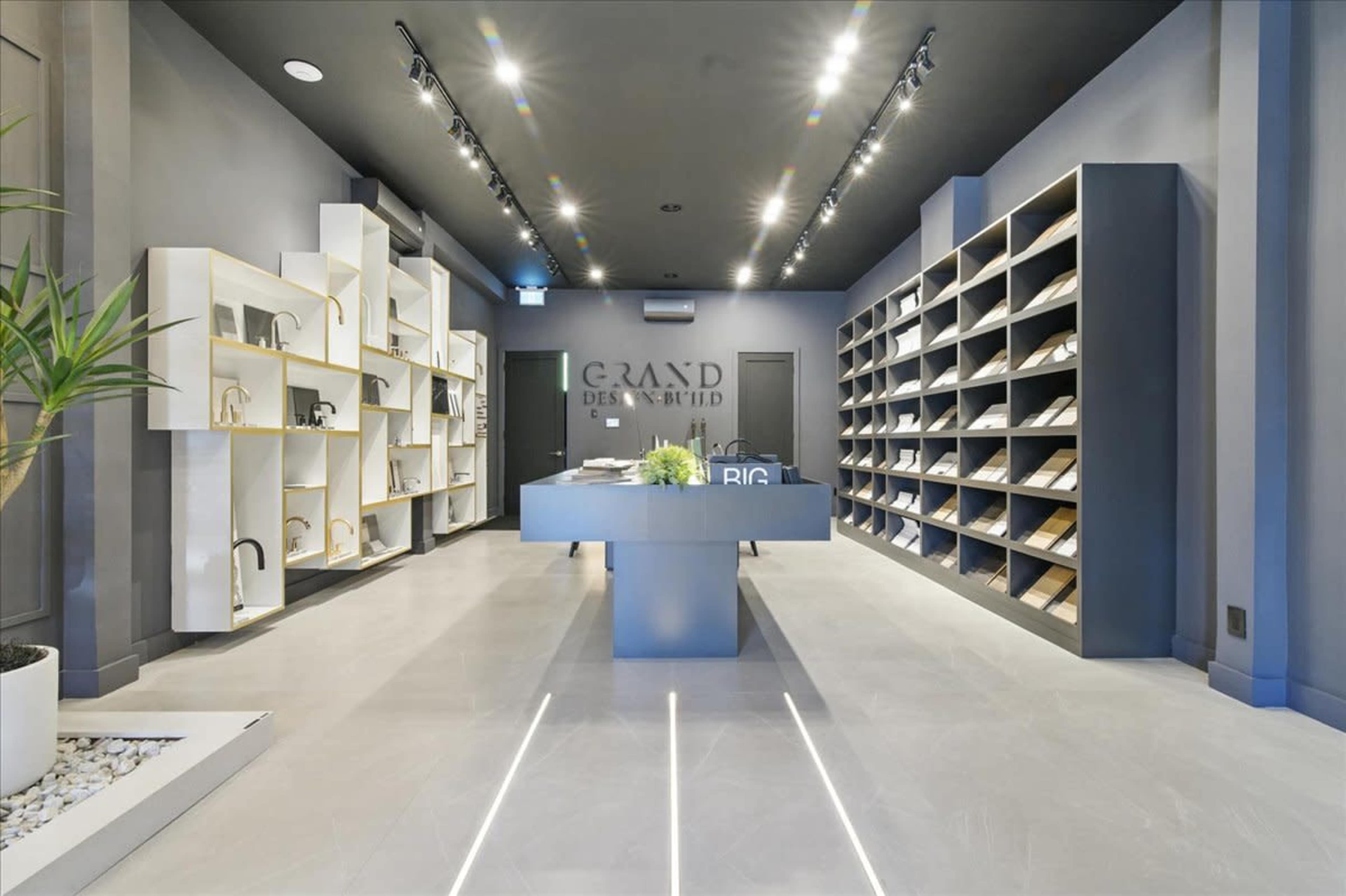 The image shows a modern interior of a design showroom featuring shelves with various display items and a central reception area.
