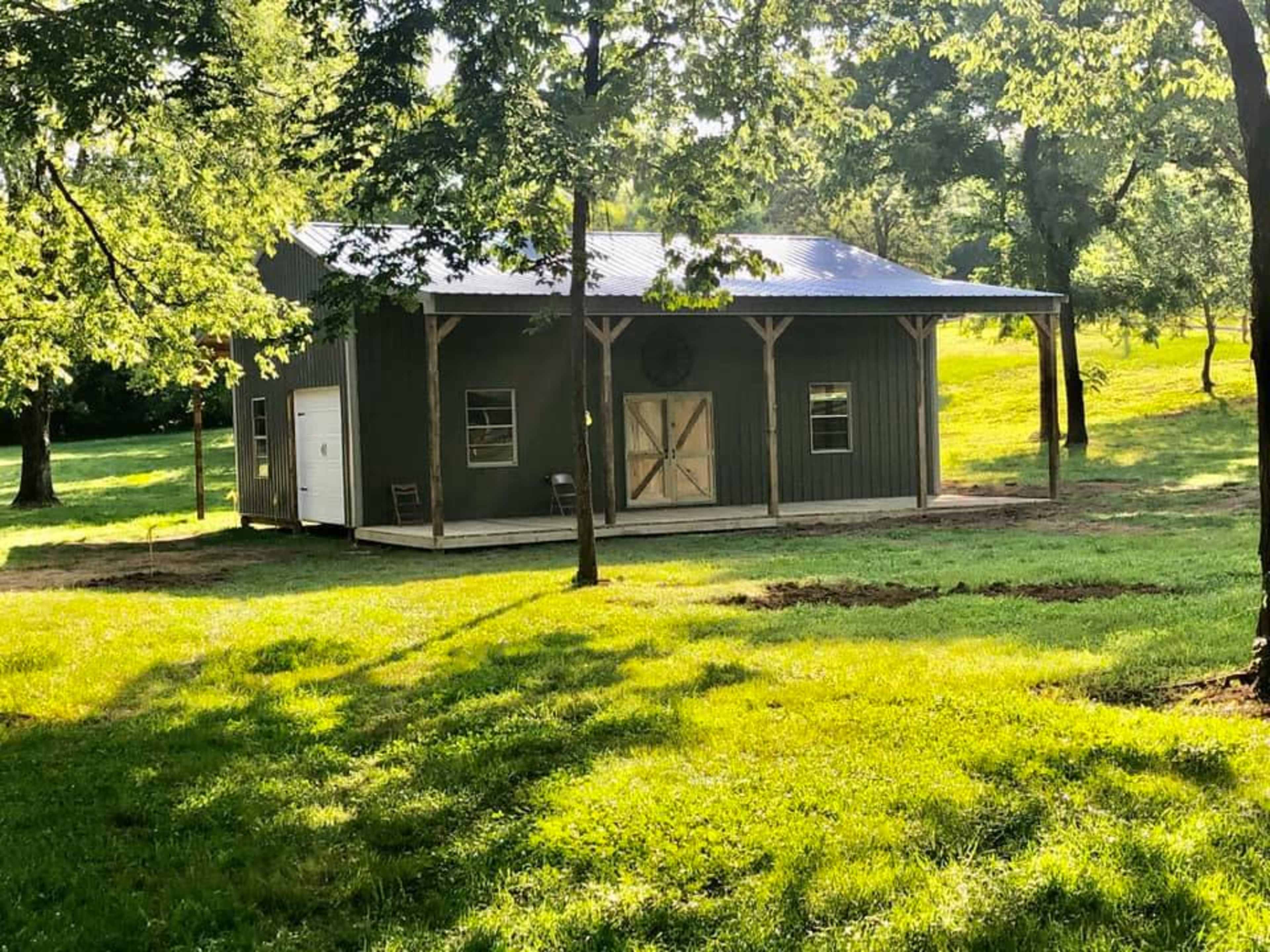 A green rustic structure with a metal roof is situated in a grassy area surrounded by trees.