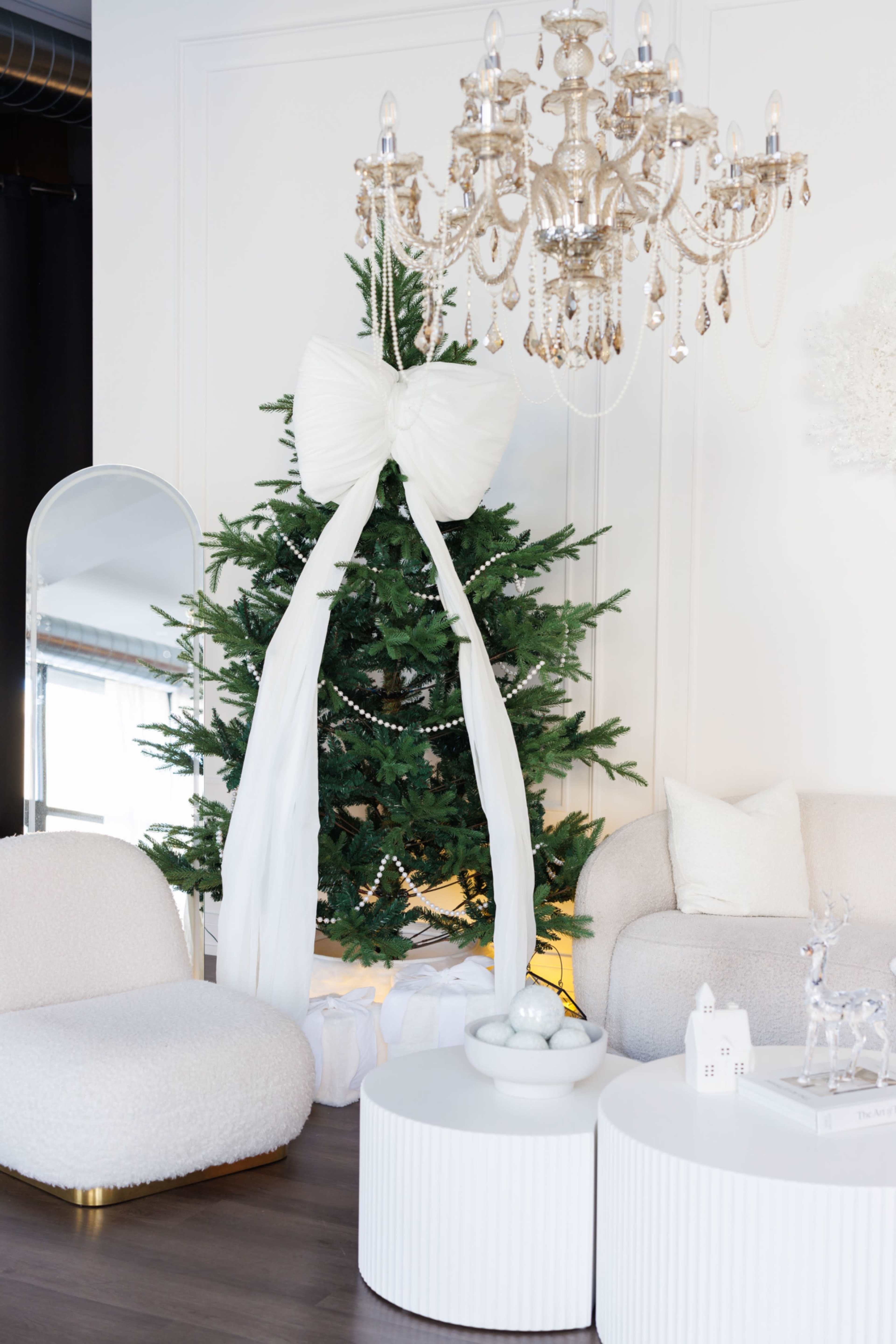 The scene features a decorated Christmas tree with a large white bow, accompanied by a chandelier, a mirror, and modern furniture in a stylish room.