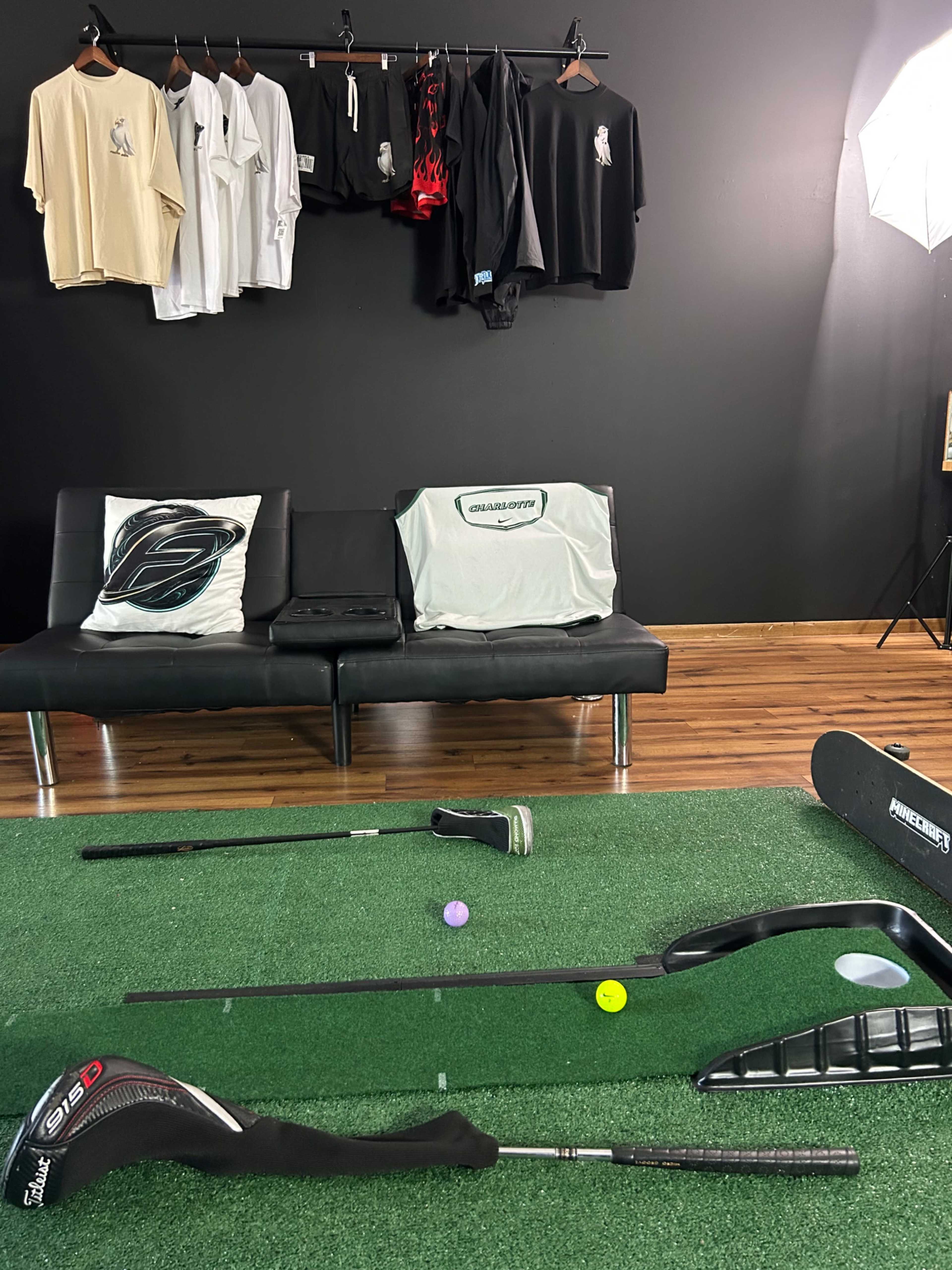 The image shows a modern indoor golf setup with a synthetic grass putting green, golf clubs, balls, and a range of golf apparel hanging on a rack in the background.