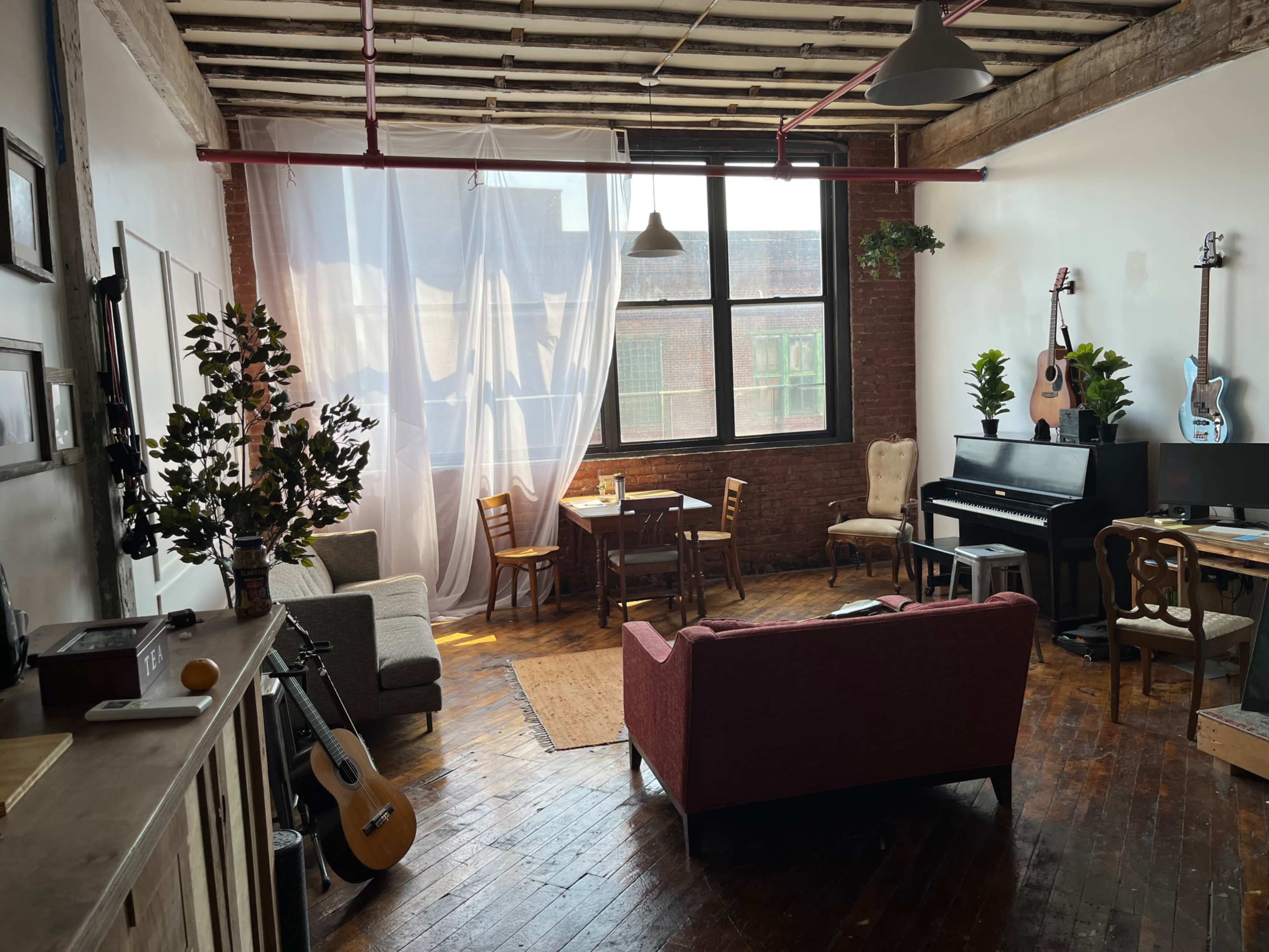 A spacious, bright living room with wooden floors, featuring a piano, a dining table, and various seating arrangements near large windows.