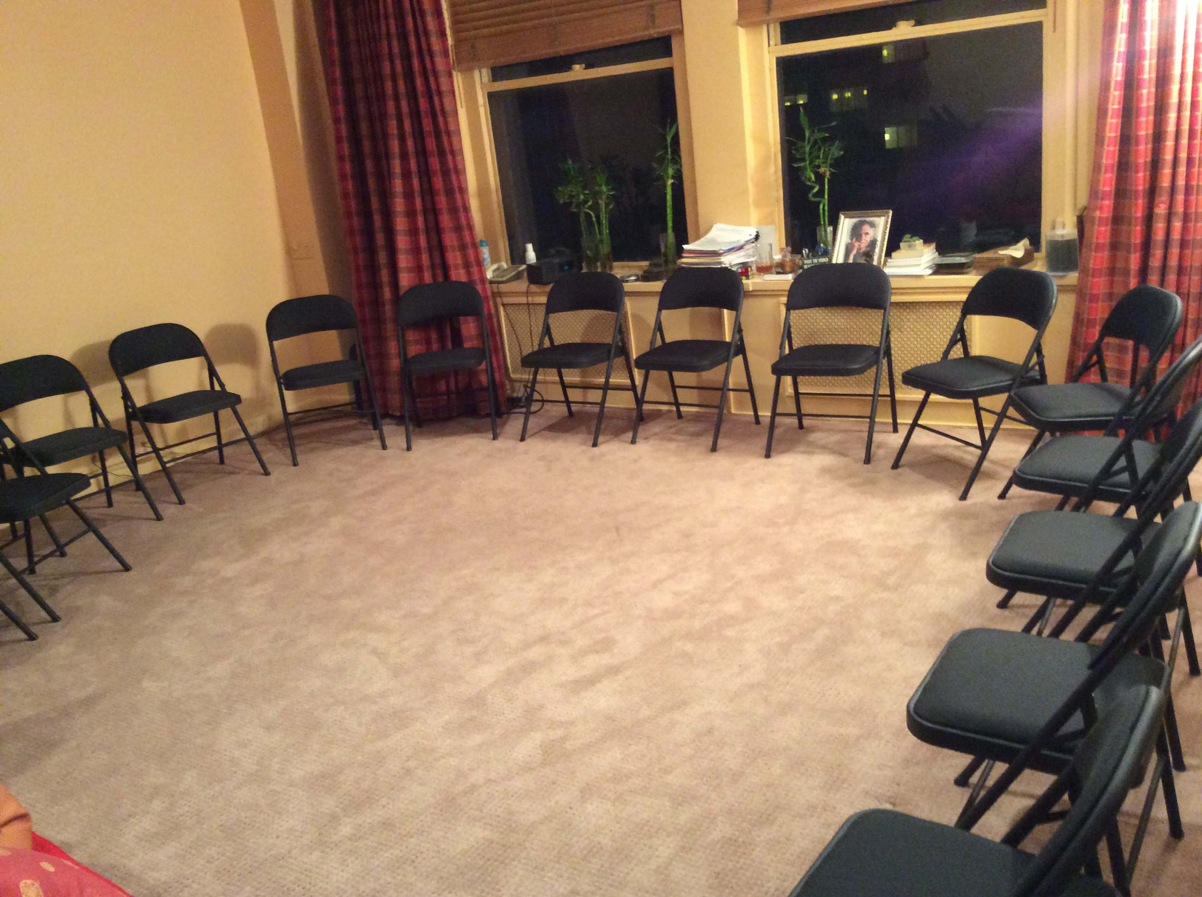 A circle of black folding chairs is arranged in a carpeted room with large windows and curtains.