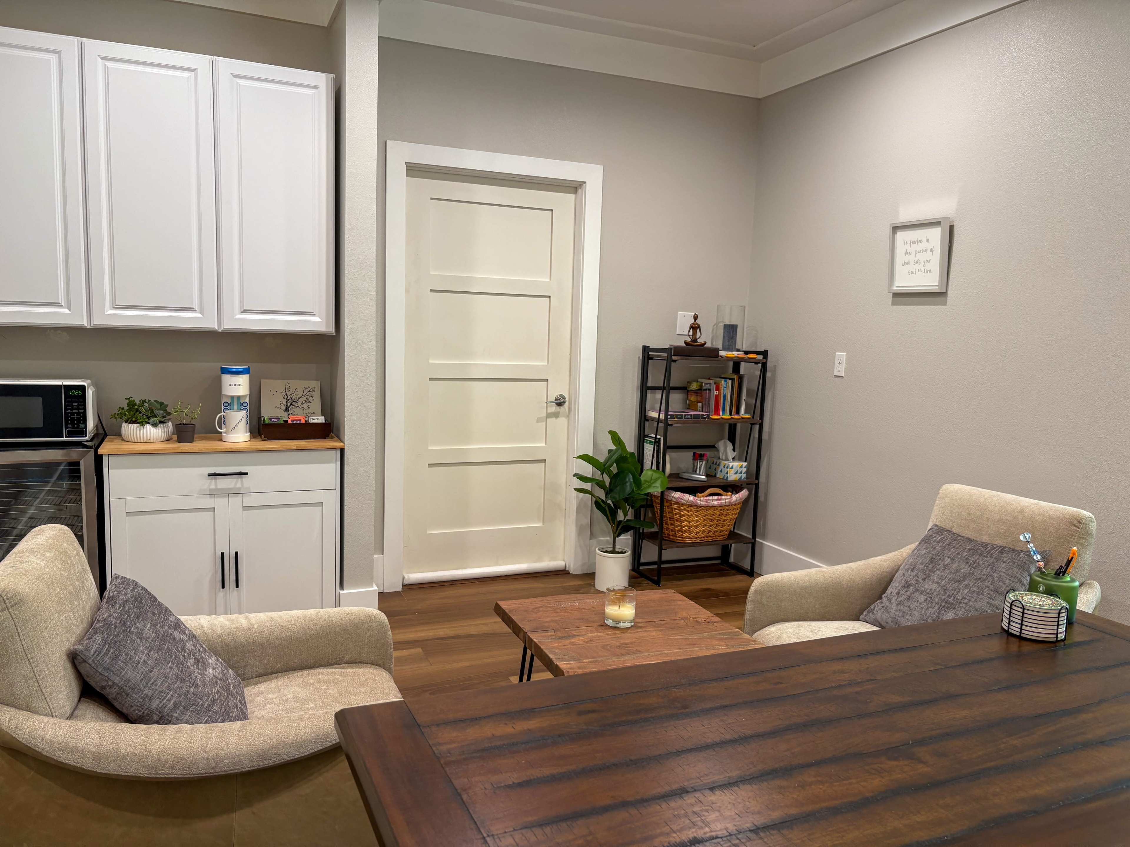 The image shows a modern office space with two armchairs, a wooden table, a bookshelf, and a kitchenette area featuring a microwave and various decorative items.