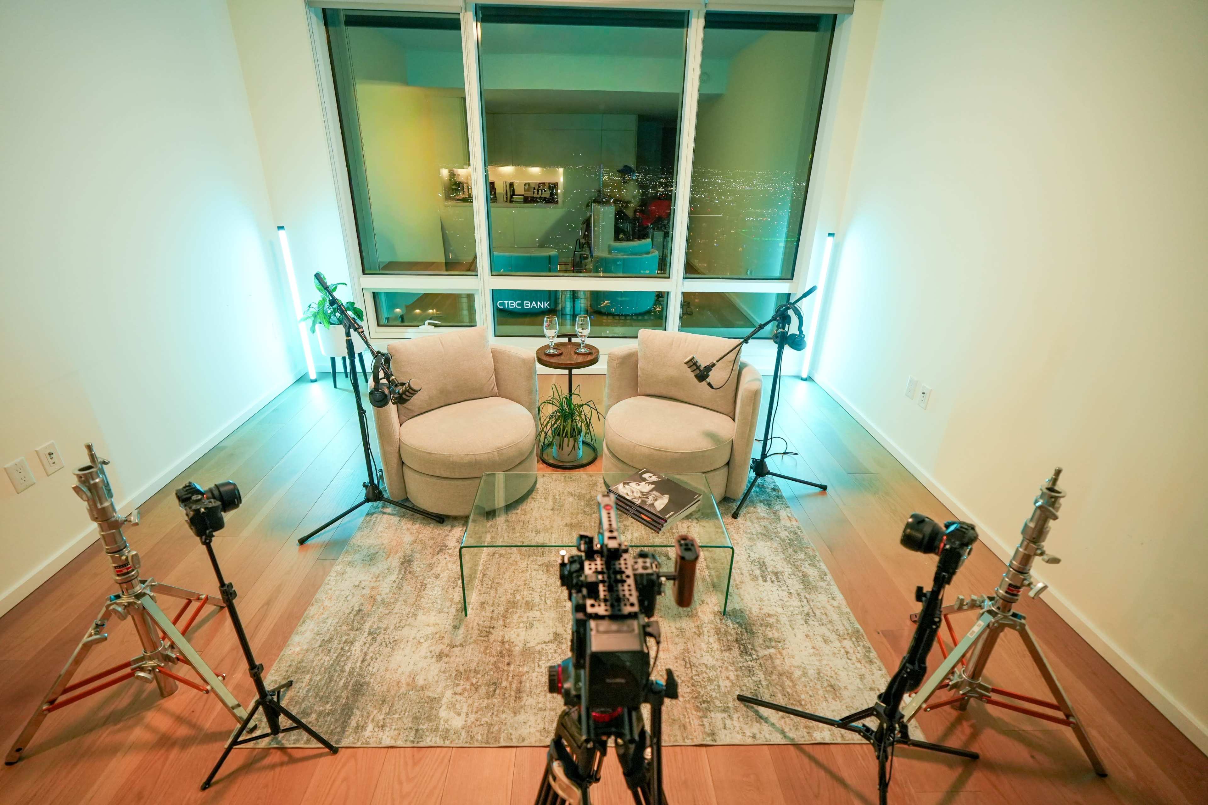 The image shows a modern indoor setup with two comfortable chairs positioned next to a plant, surrounded by camera equipment and lighting, set against a large window with a city view.
