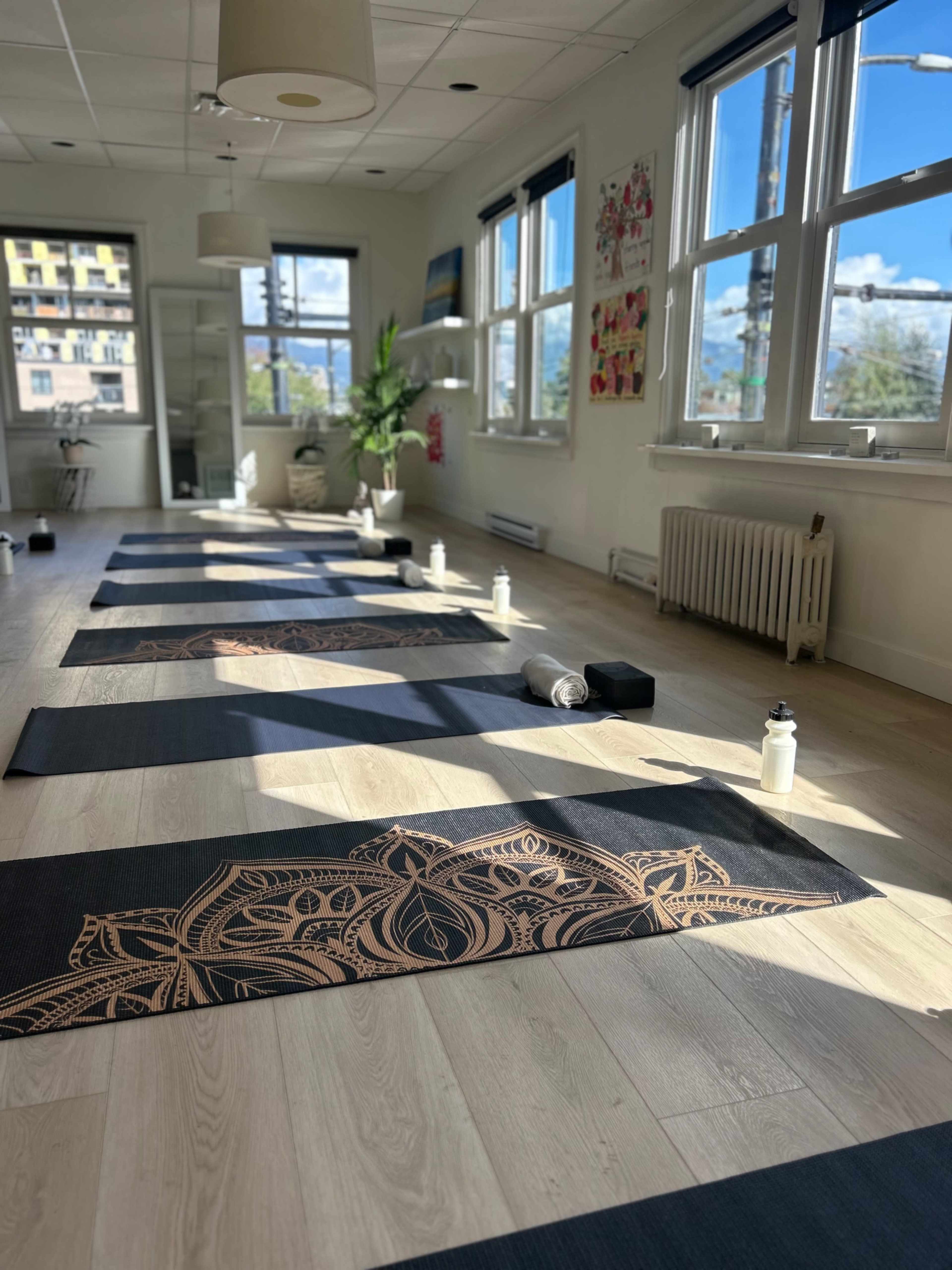 The image shows a yoga studio with several mats arranged neatly on a wooden floor, featuring water bottles and blocks on each mat, illuminated by natural light from large windows.