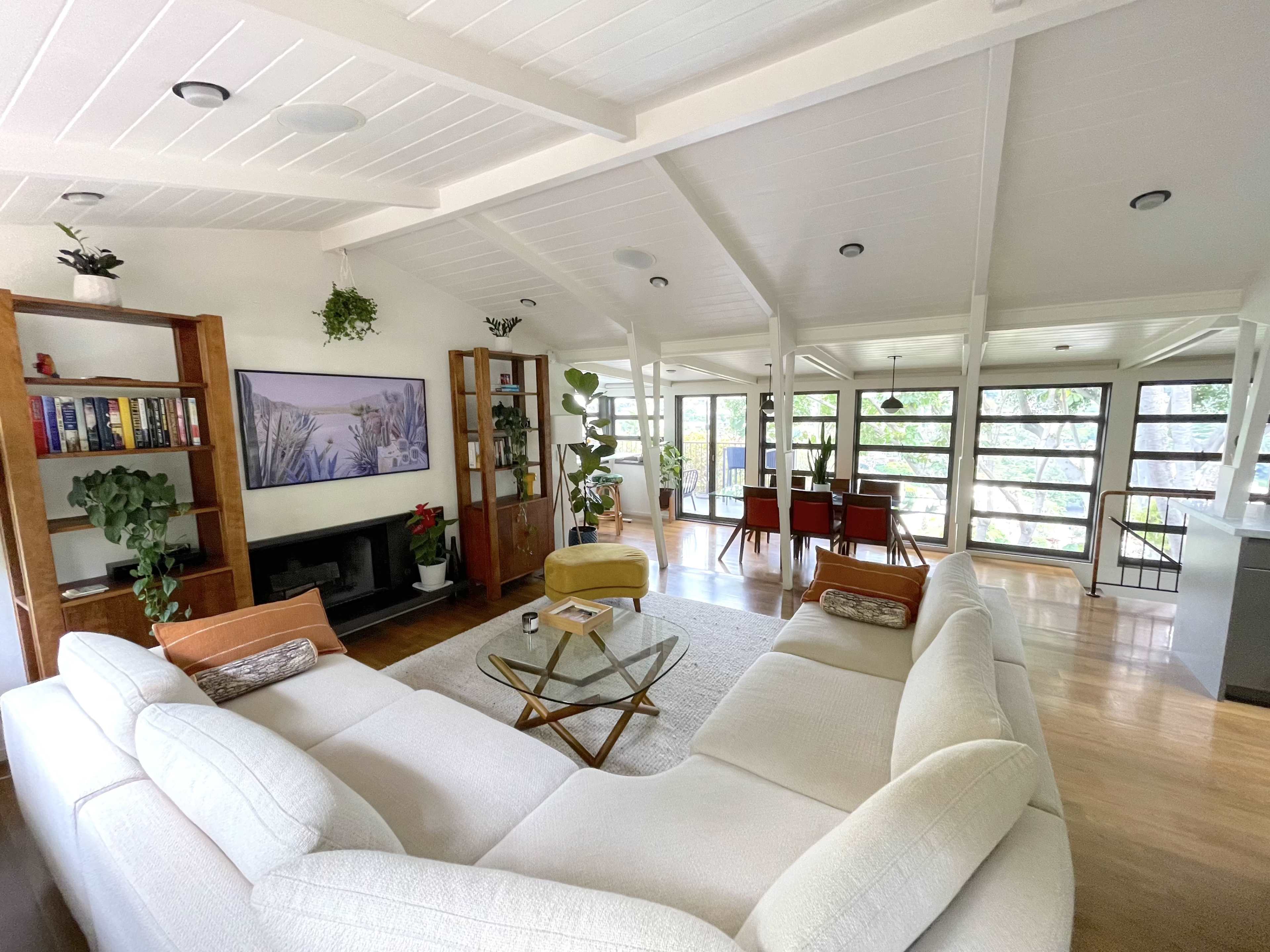 A spacious living area features a white sectional sofa, a glass coffee table, and large windows overlooking greenery.