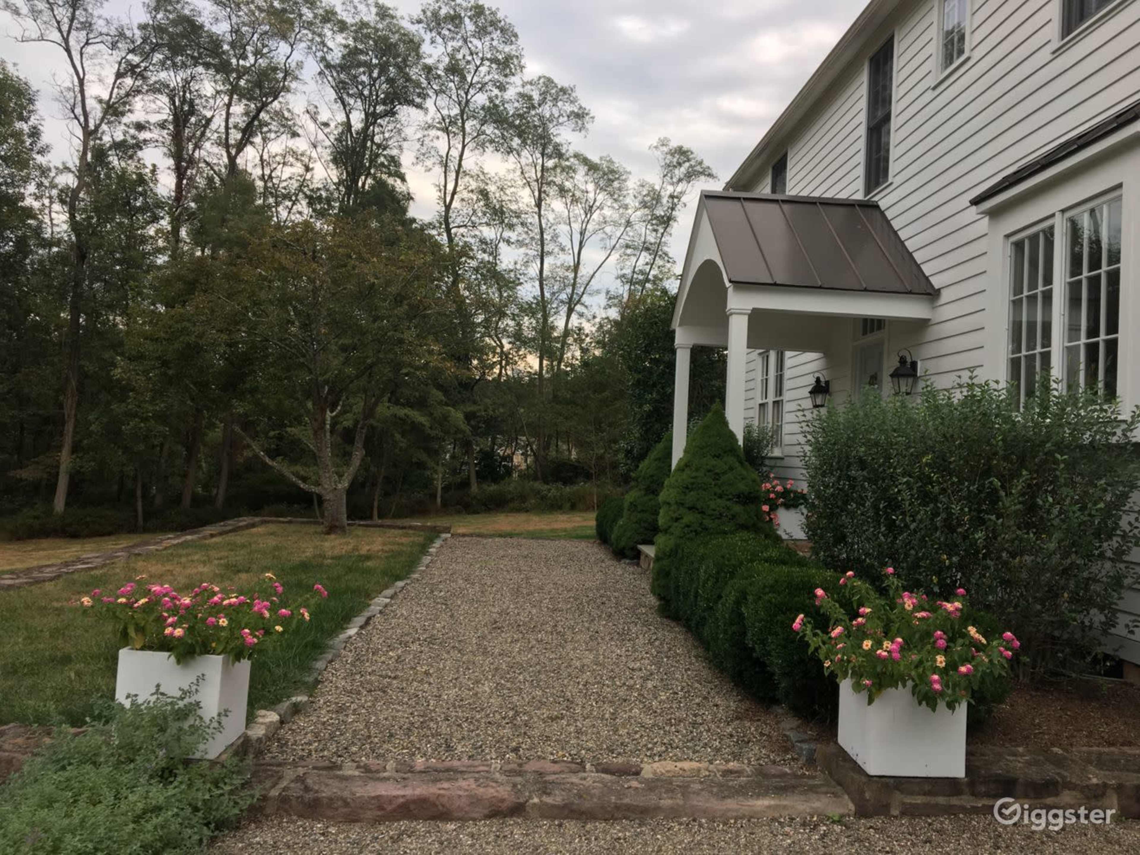 A gravel pathway lined with planters of pink flowers leads to the entrance of a white house with a covered porch.