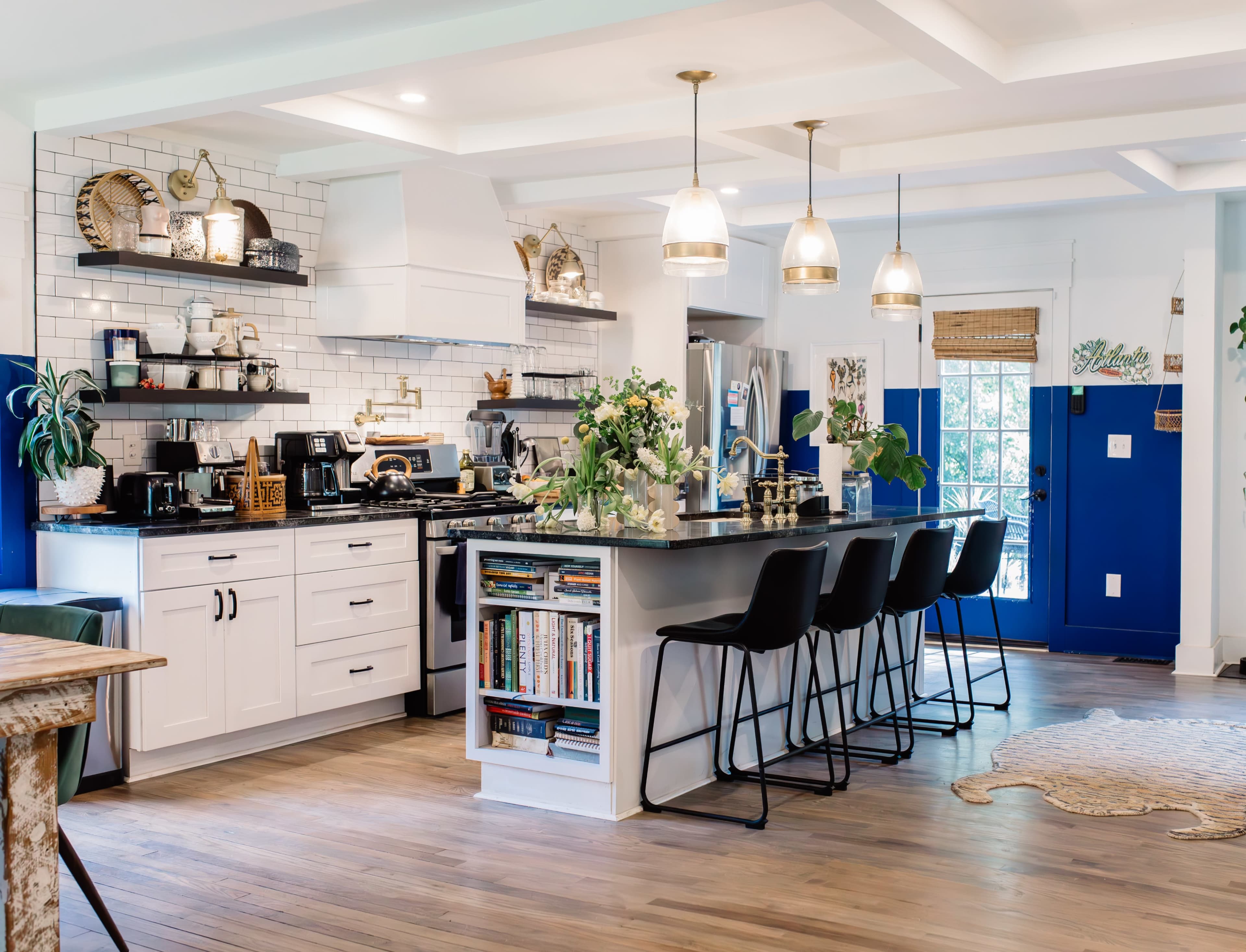 The image shows a modern kitchen with white cabinetry, a black countertop, and bar stools, featuring open shelving and decorative plants.