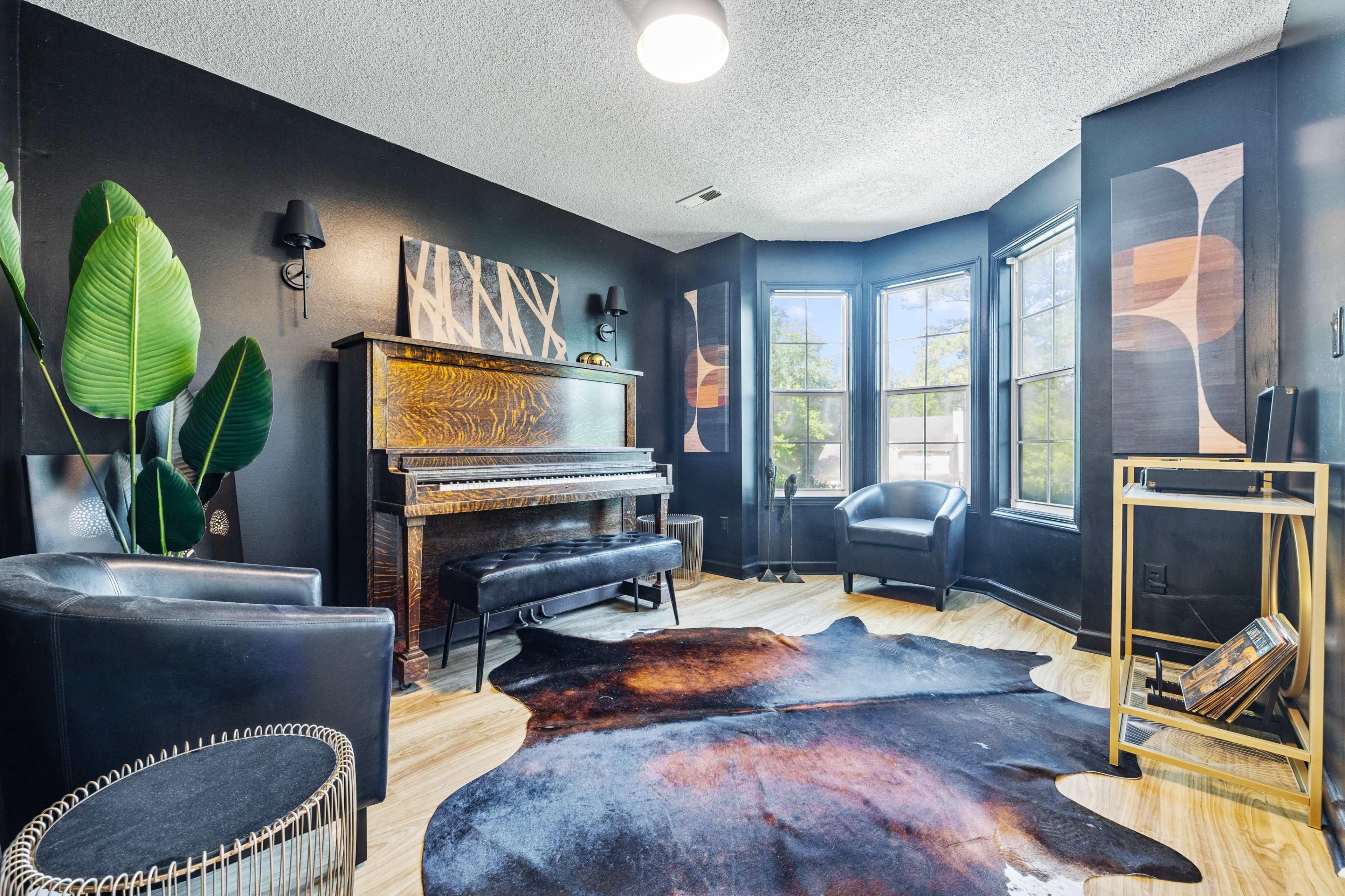 A corner of a room features a piano, two black chairs, a cowhide rug, and large windows with decorative wall art.
