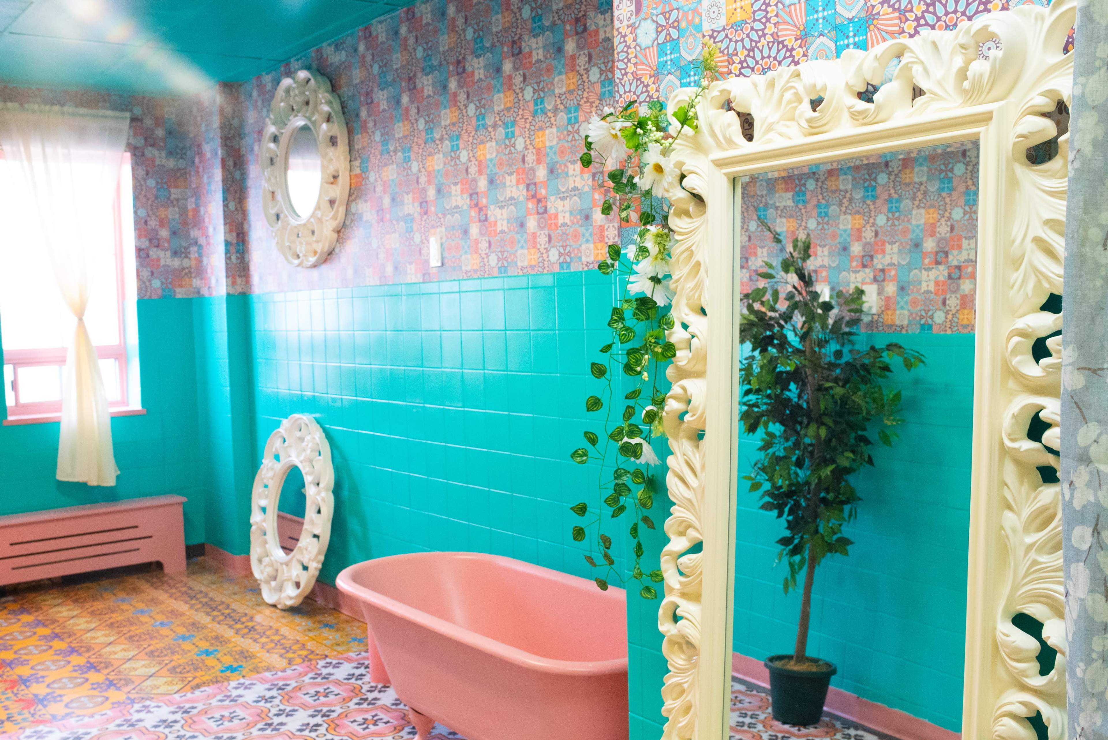 A colorful bathroom featuring a pink bathtub, ornate mirrors, and vibrant patterned tiles on the walls and floor.