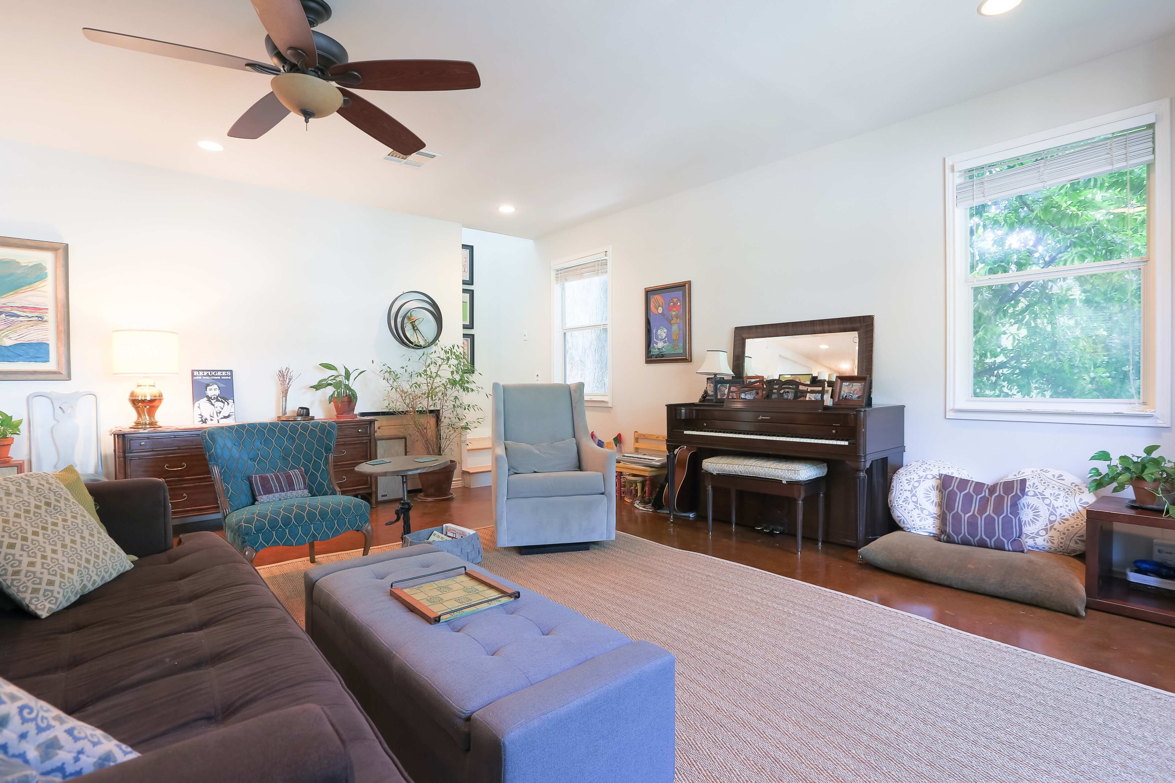 A bright living room features a brown sofa, a gray armchair, a piano, and potted plants, with natural light streaming through the windows.