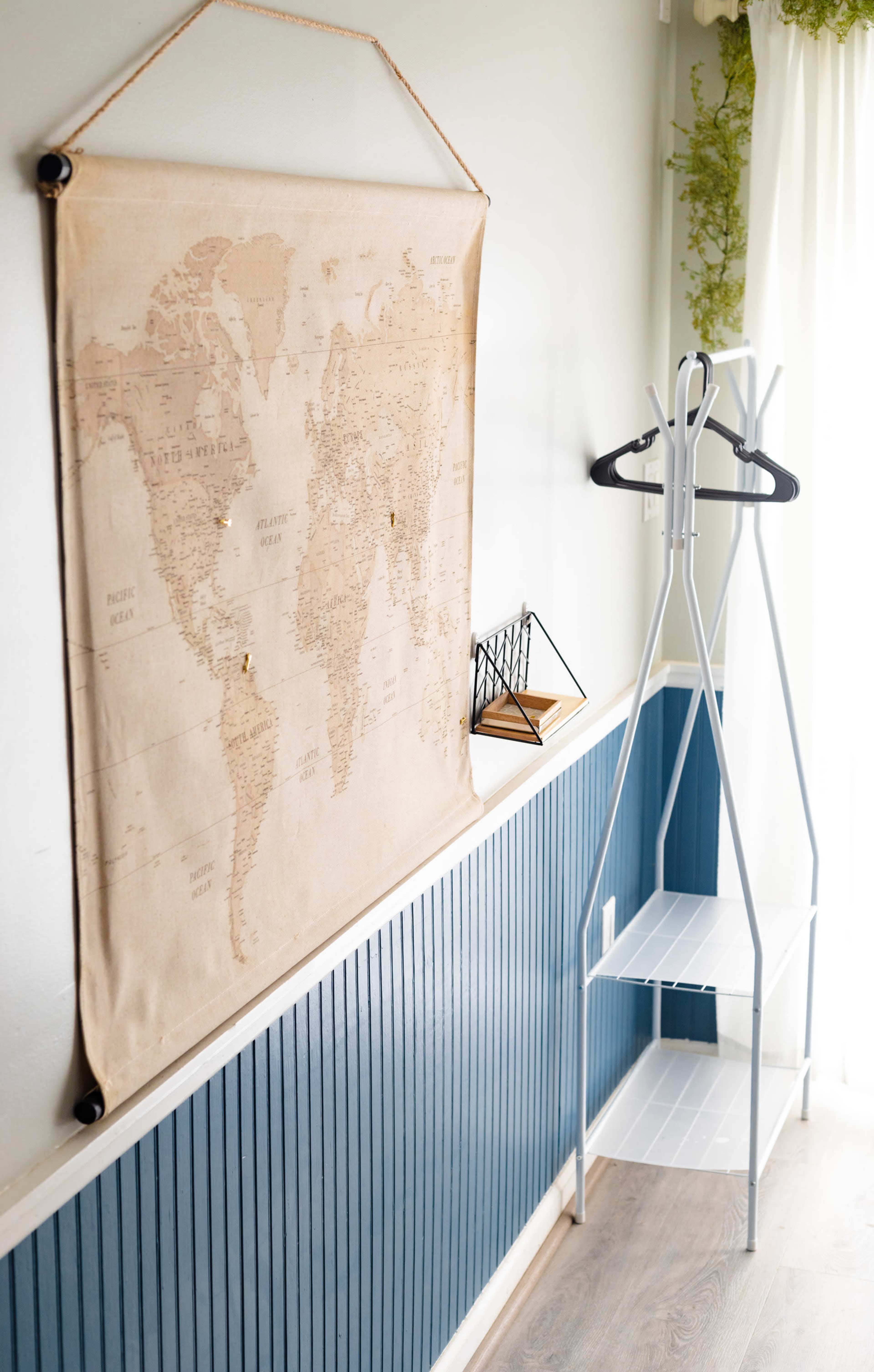 A wall with a world map tapestry, a geometric decorative item on a ledge, and a simple white clothing rack in a room with blue wainscoting.